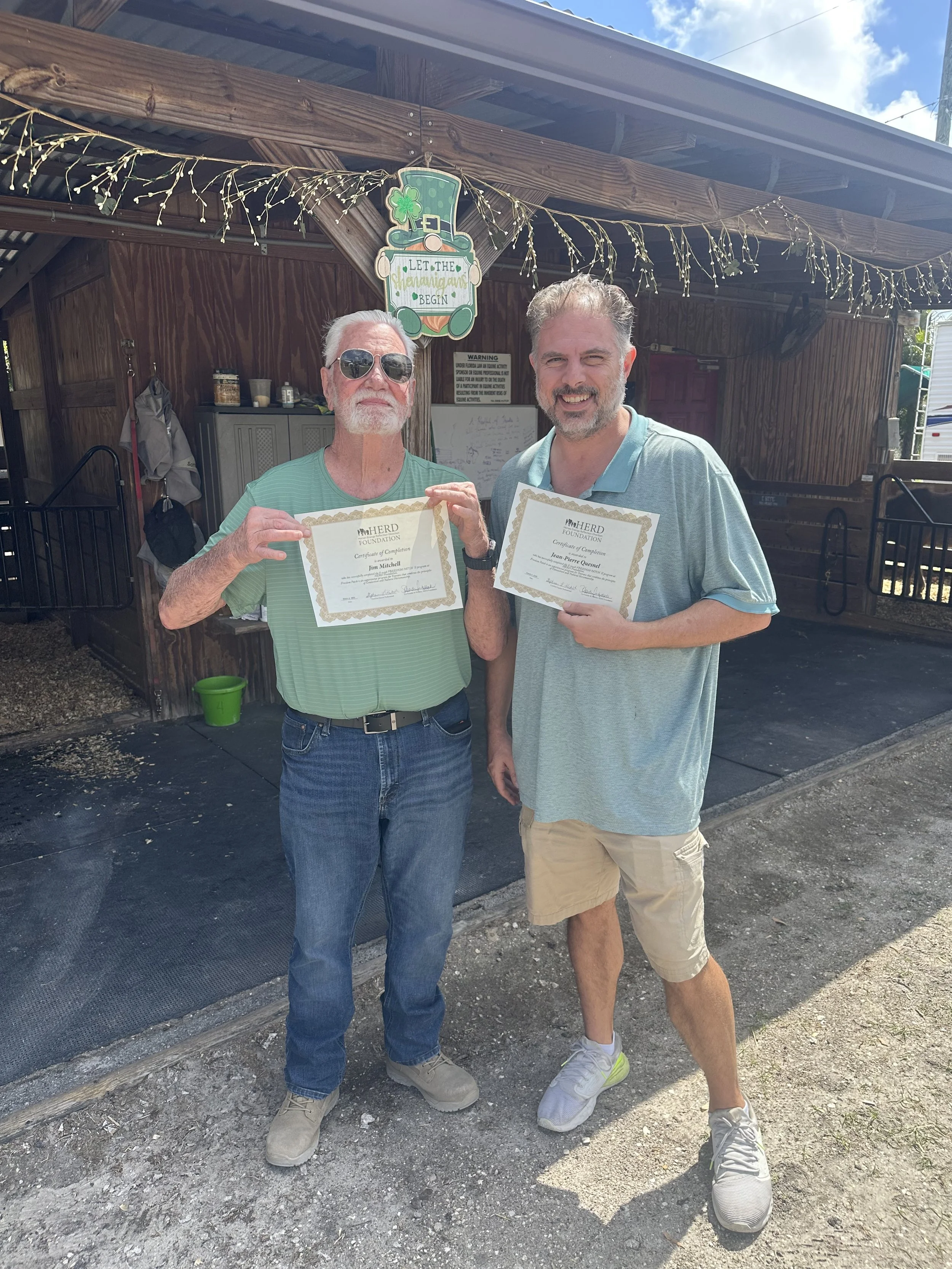 Two men standing outside in front of a wooden structure, holding certificates, with a St. Patrick's Day sign and string lights above them. One man is wearing sunglasses and a green striped shirt, the other is smiling and wearing a light blue shirt an