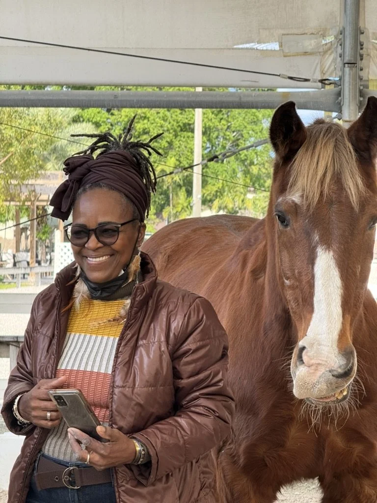 A smiling woman with glasses, wearing a brown jacket, colorful striped shirt, and a headwrap, standing next to a brown horse with a white stripe on its face, under a tent structure outdoors with green trees and a cloudy sky in the background.