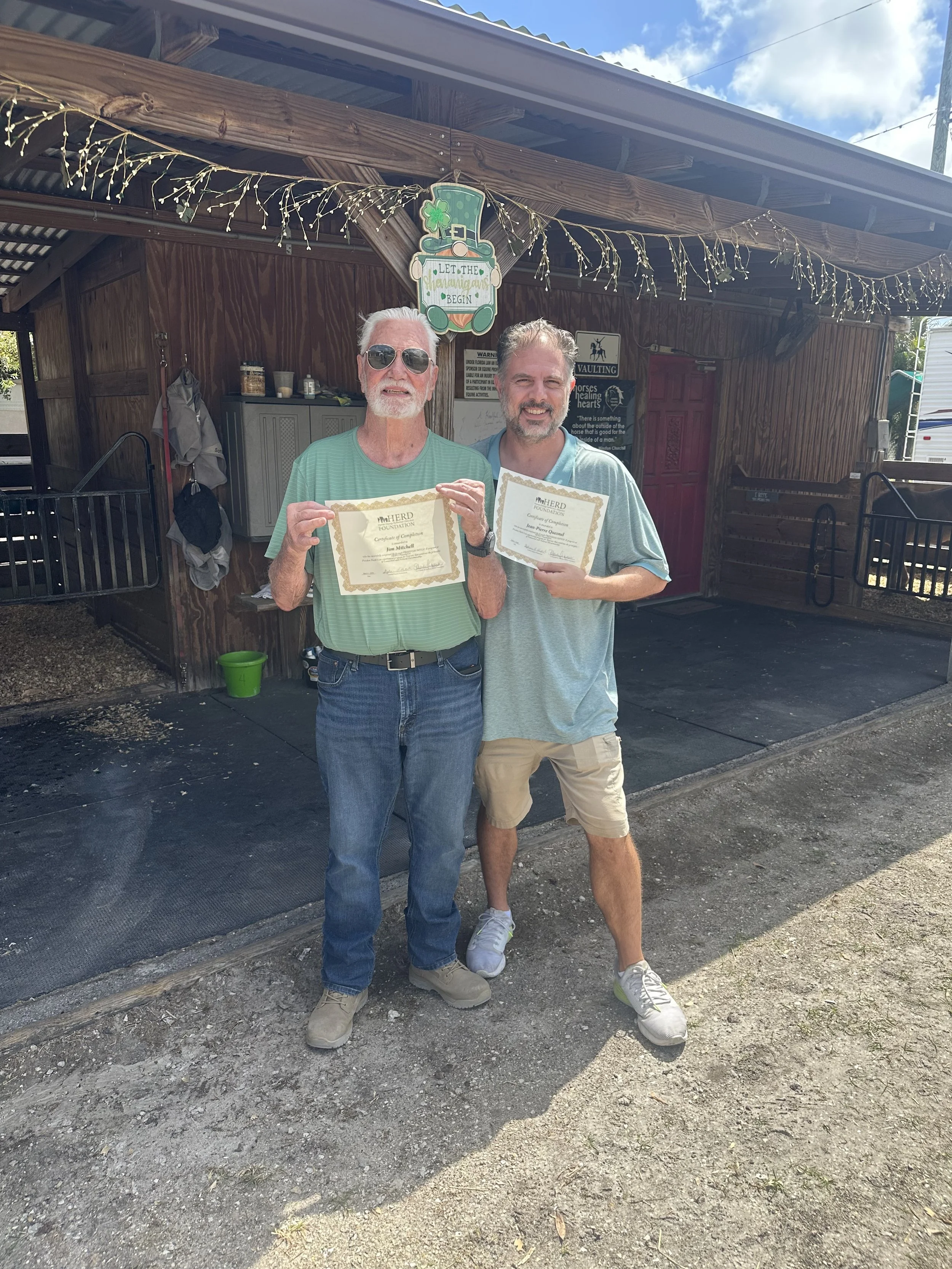 Two men standing outdoors in front of a wooden shed, holding certificates and smiling. The older man on the left is wearing sunglasses and a green striped shirt, the younger man on the right is wearing a light blue shirt and beige shorts. The shed is decorated with fairy lights and has a sign that reads "Let the shenanigans begin" with festive decorations.