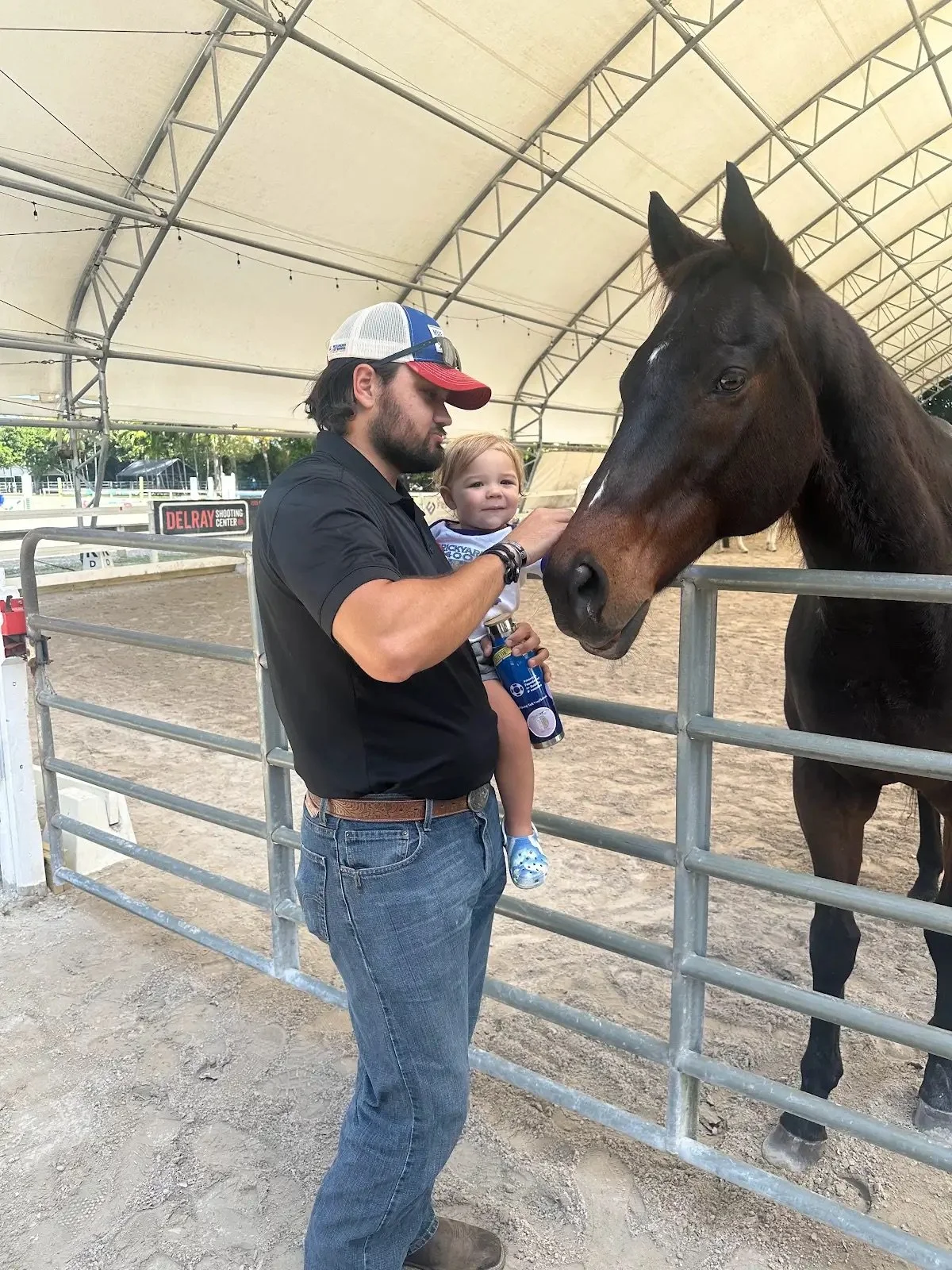 A man in a black shirt and jeans holding a young girl in a white shirt with blue Crocs, feeding a brown horse through a metal fence inside a covered riding arena.