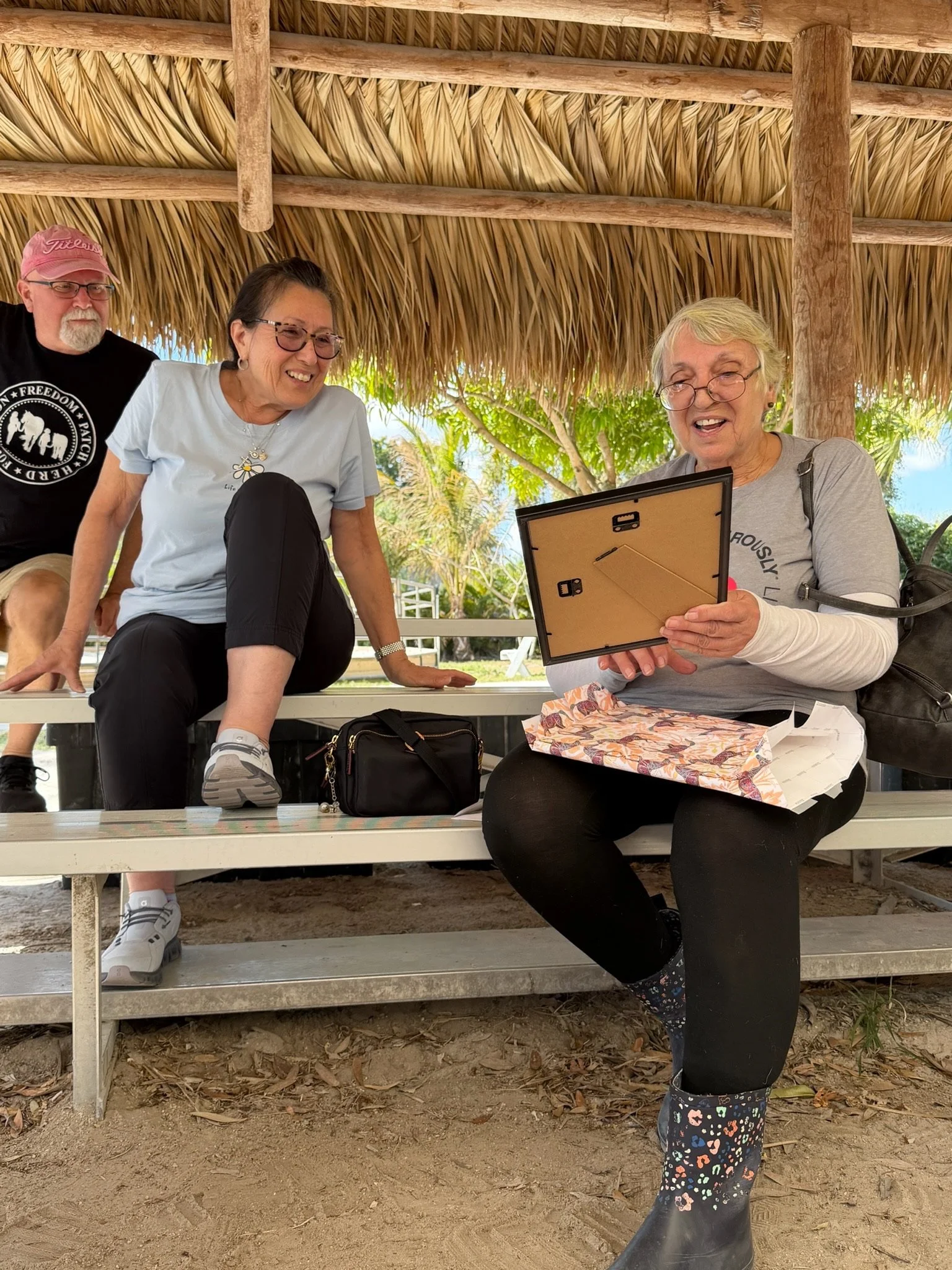 Four people sitting and standing under a thatched roof structure, with two women sitting on the benches and a man standing behind them, all smiling and looking at a photo frame held by one woman. The scene outdoors with trees and sunlight in the back