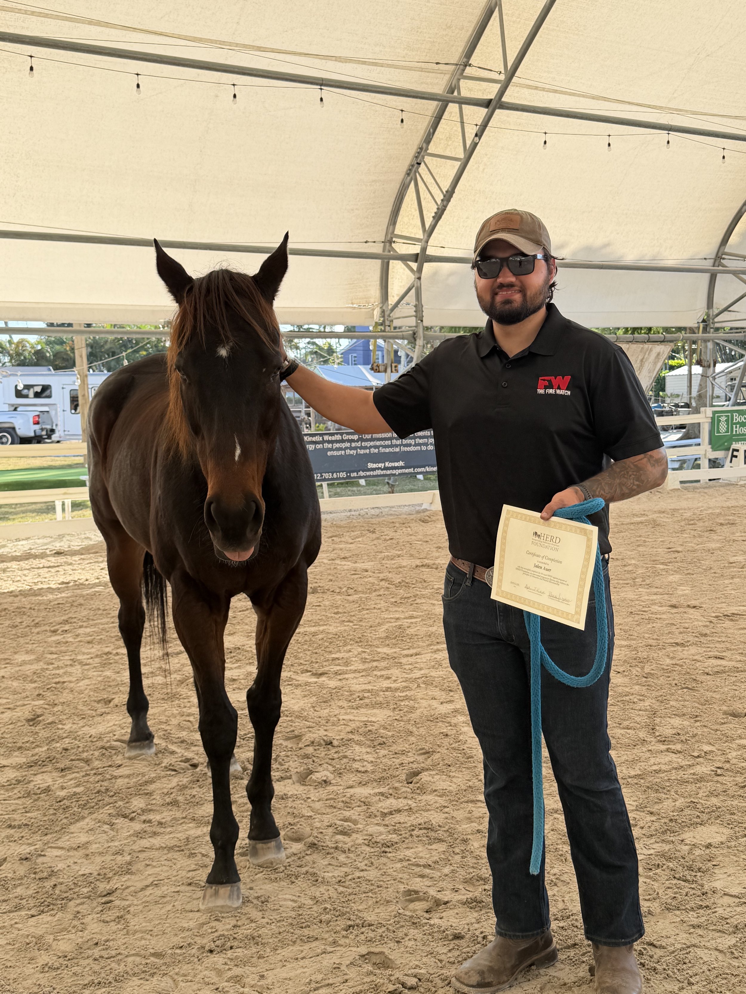 A man in a black polo shirt and jeans, wearing sunglasses and a cap, holding a certificate and a blue lead rope, rests his hand on a brown horse inside a covered riding arena.