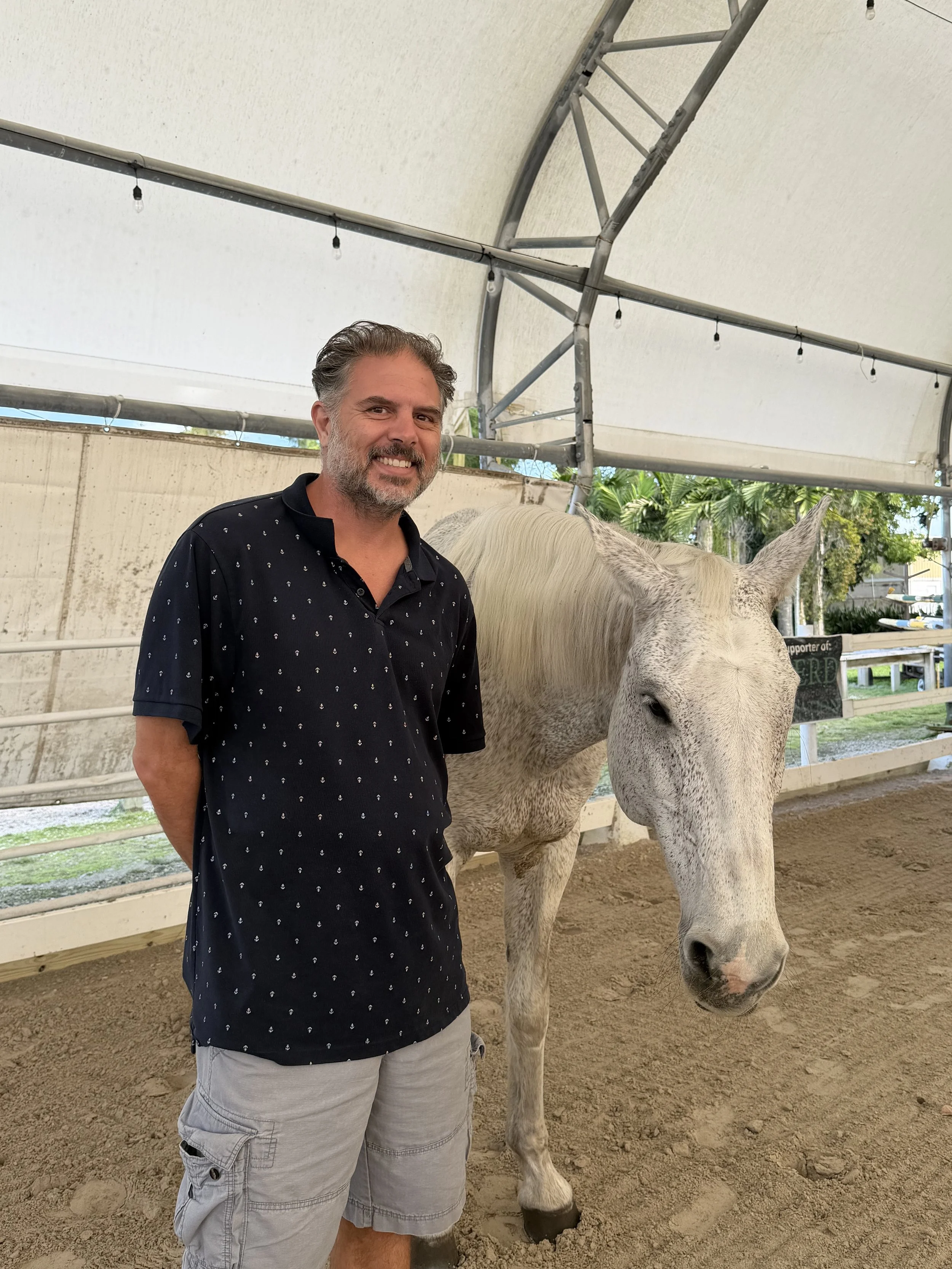 A man standing next to a white horse inside a covered outdoor arena with a dirt floor and fencing in the background.