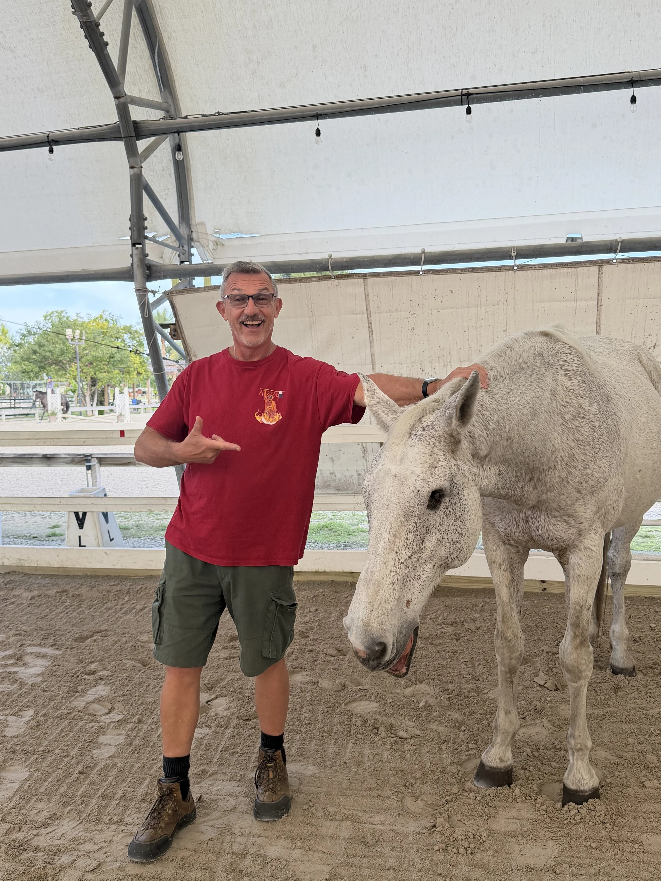 A smiling man in a red shirt and khaki shorts petting a white horse inside a covered outdoor riding arena.