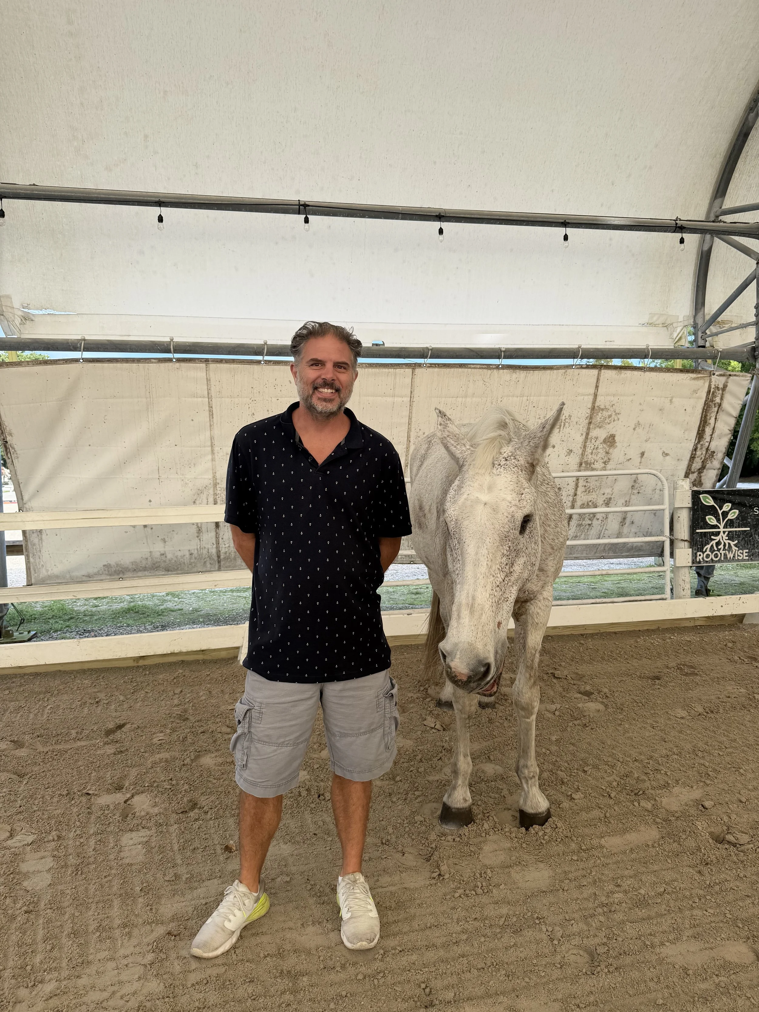 A man standing next to a white horse inside a covered outdoor arena.