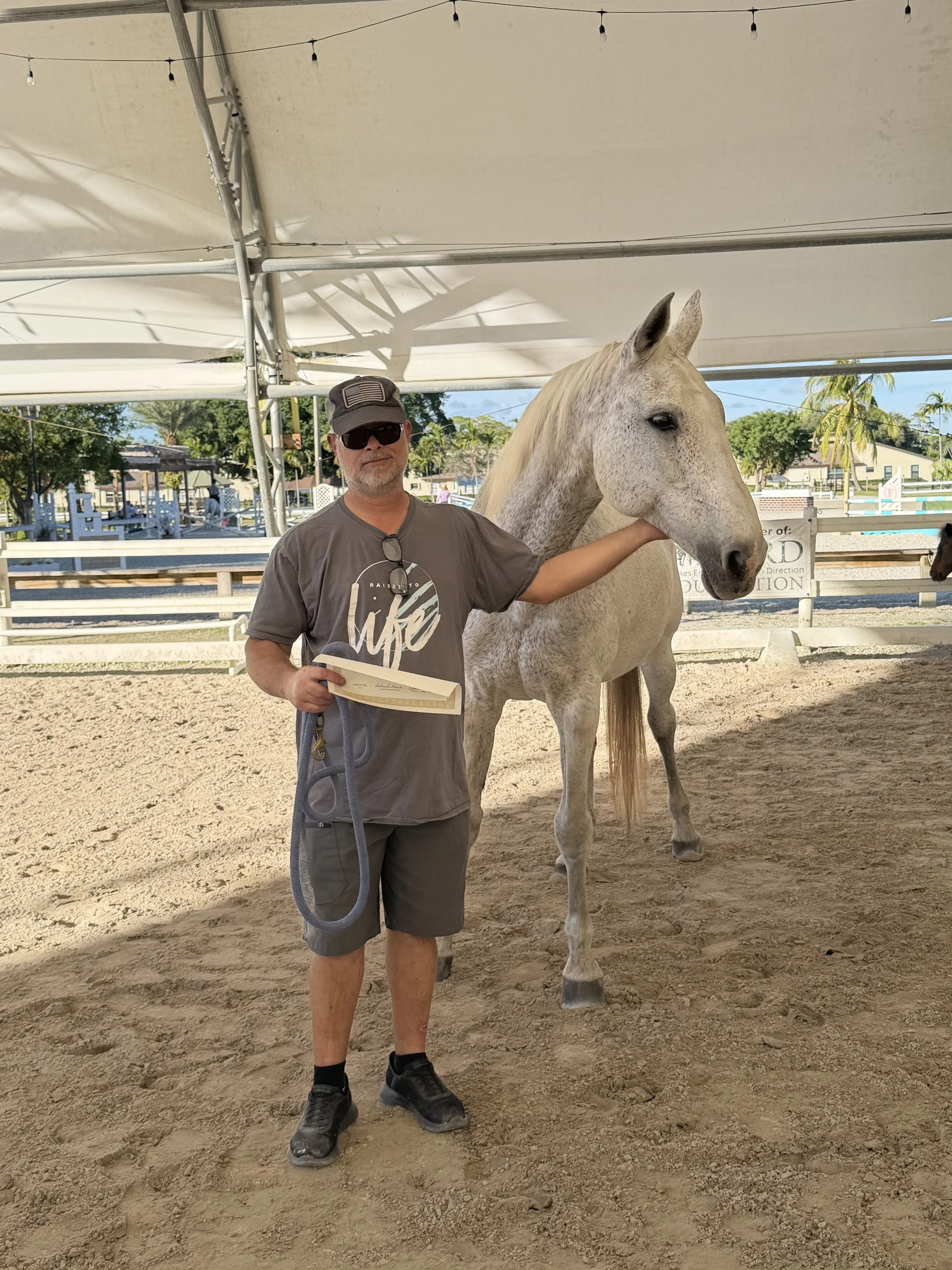 A man wearing sunglasses, a cap, a gray T-shirt, and shorts standing next to a light-colored horse under a large tent, with palm trees and a riding arena visible in the background.