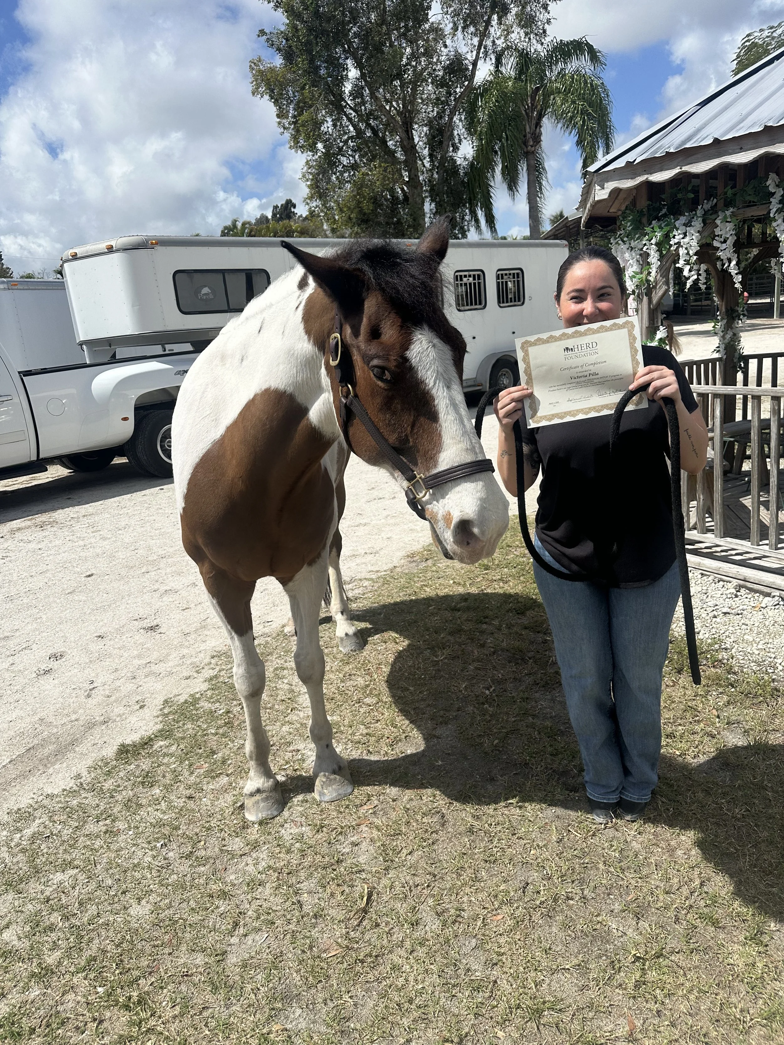 A woman holding a certificate standing next to a brown and white horse outdoors near a white trailer and a wooden structure with white flowers and palm trees in the background.