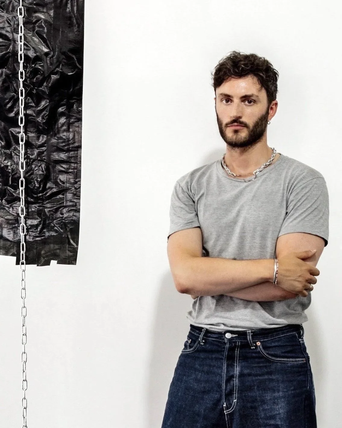 A young man with short curly hair and a beard, wearing a light gray t-shirt, jeans, and silver jewelry, standing with arms crossed against a plain white wall, with a black textured curtain and chain on the side.
