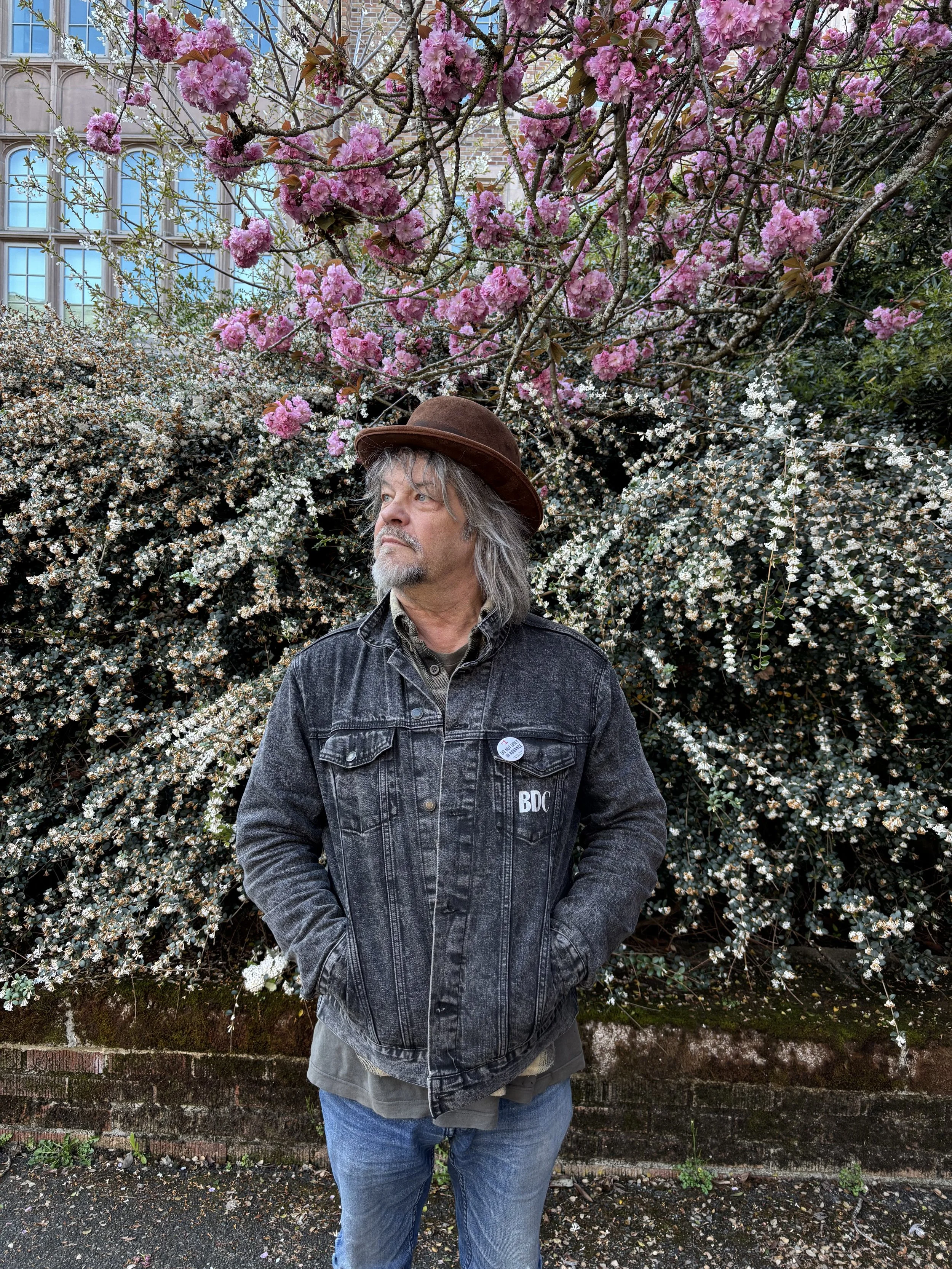 A man with gray hair and a beard wearing a brown hat, a black denim jacket with patches, and blue jeans stands in front of flowering bushes with pink and white blossoms.