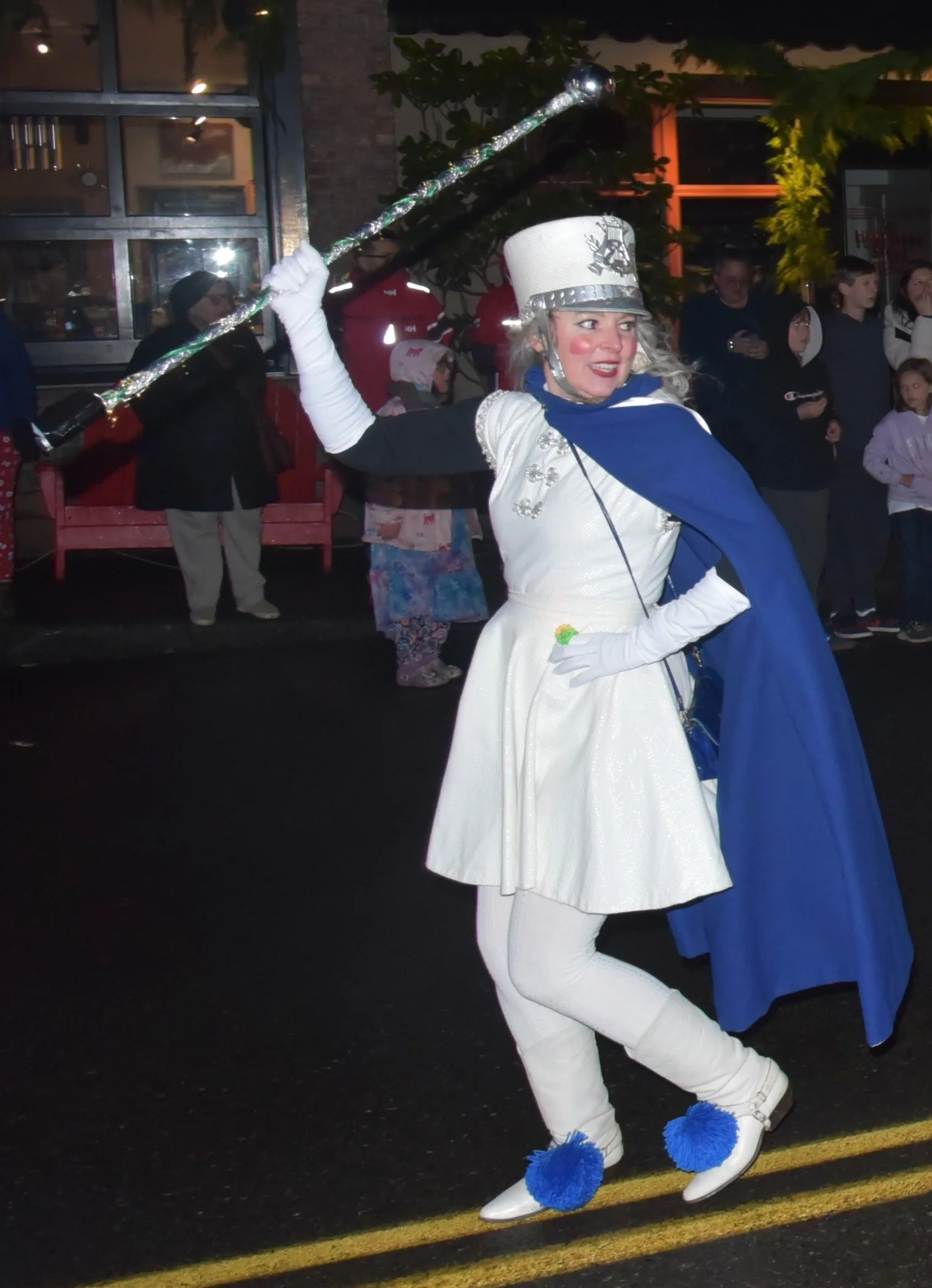 Woman dressed as a snow queen or princess holding a glittery staff at a parade at night, surrounded by onlookers.