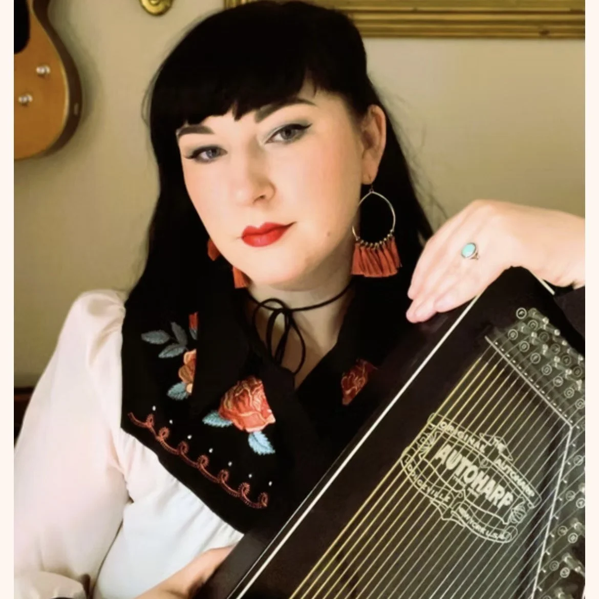 A woman with black hair, red lipstick, and traditional embroidered blouse holding a cigar box guitar.