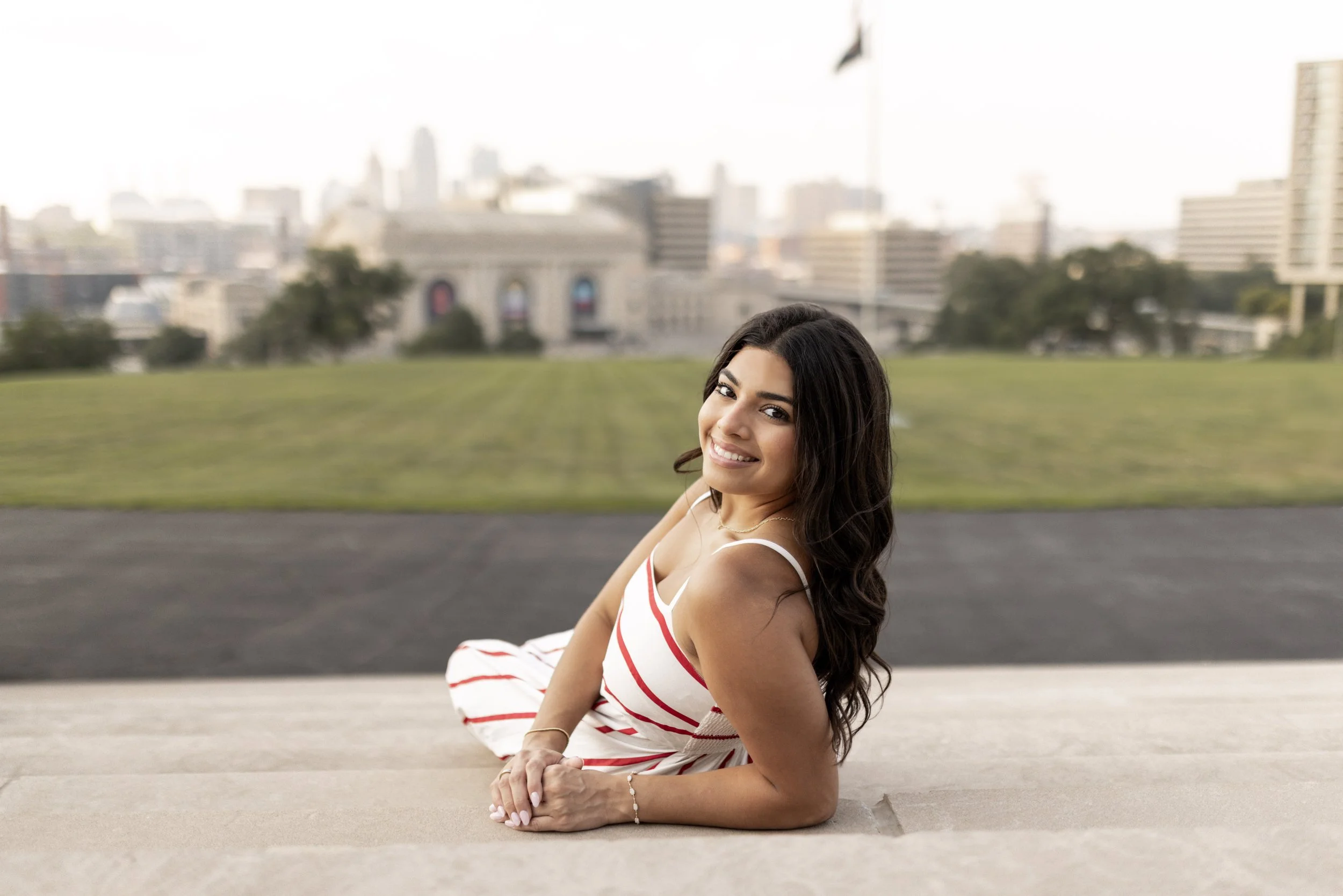 Smiling woman with long dark hair lying on the ground in an urban park, wearing a white dress with red stripes, with city buildings in the background. Kansas City Senior Photographer