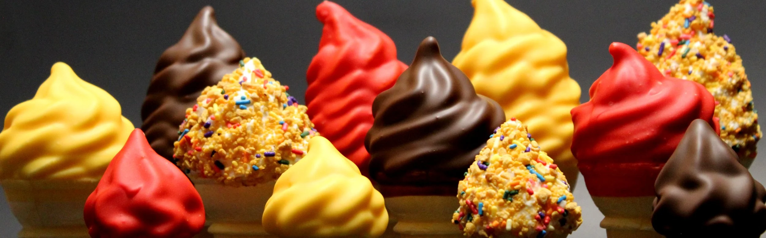 Assorted colorful ice cream cones with various toppings, including sprinkles and chocolate coating, on a dark background.