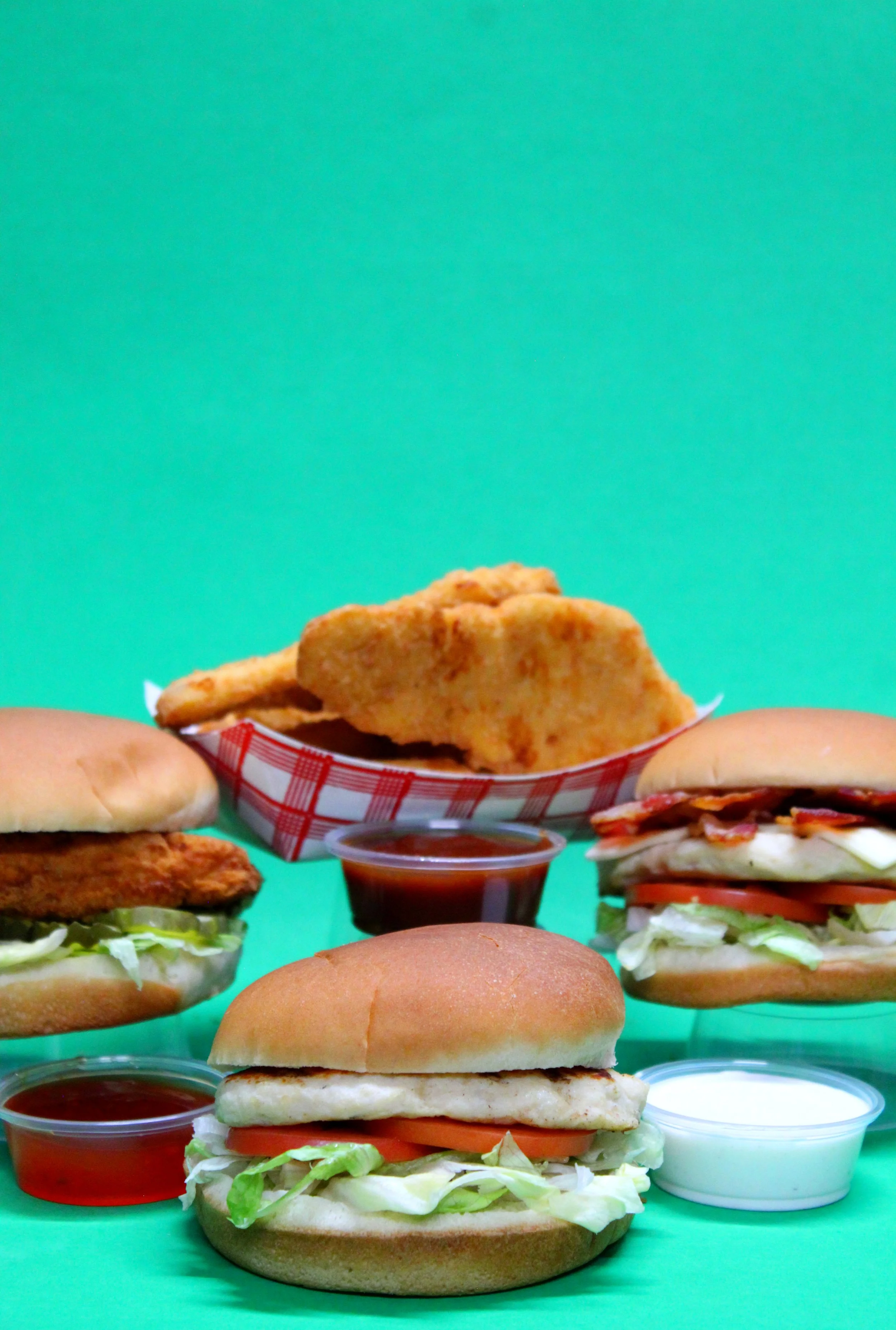 Assorted fast food, including chicken sandwiches, fried chicken tenders, and dipping sauces placed in front of a bright green background.