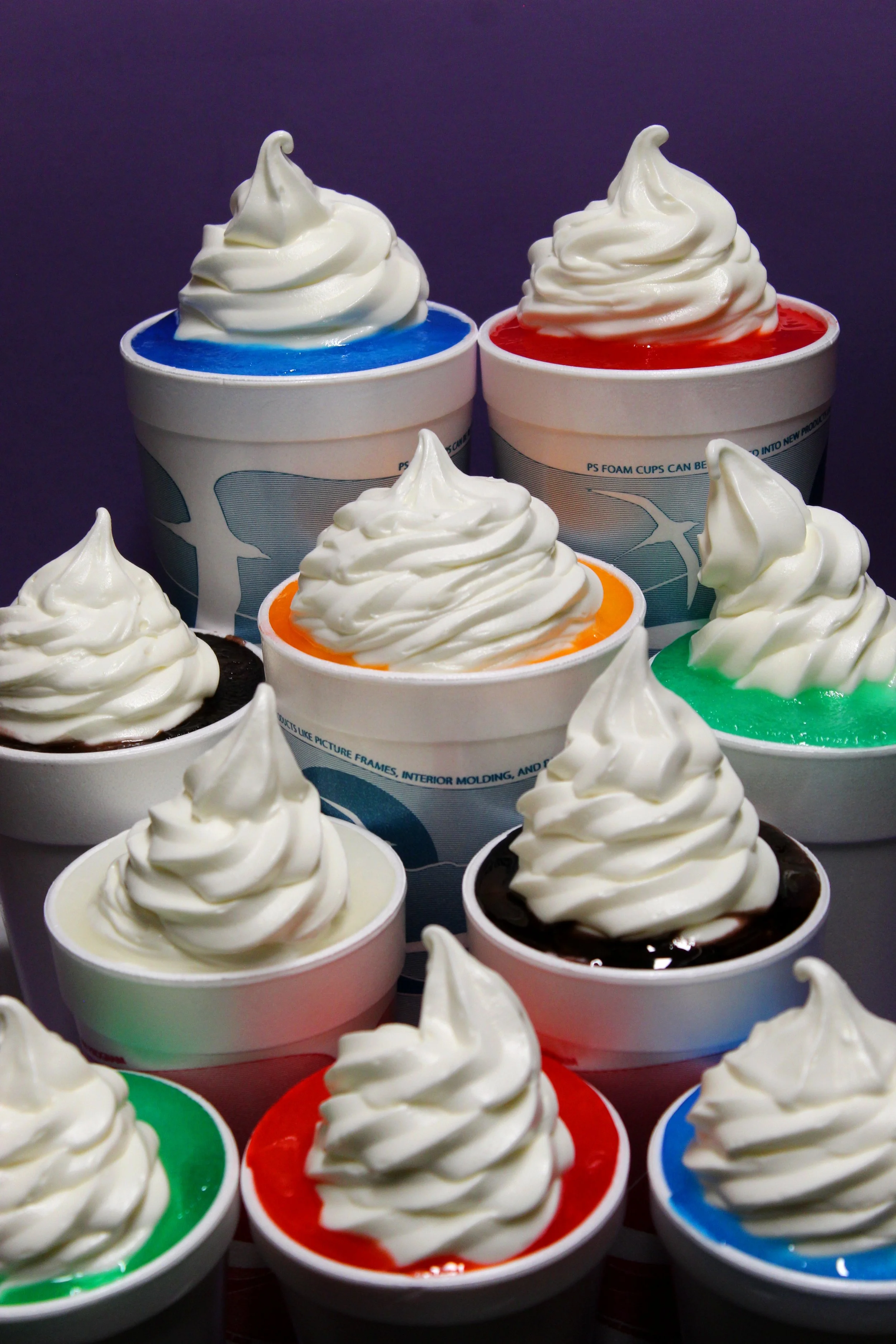 Multiple cups of colorful soft serve ice cream with swirled peaks, arranged in a pyramid-like display against a dark background.