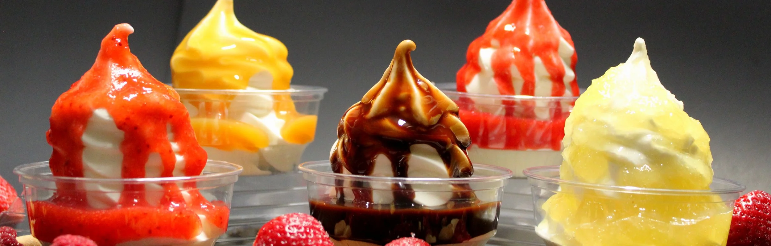Various soft-serve ice creams topped with fruit sauces, chocolate, and drizzled in caramel, displayed in clear cups with strawberries in front.