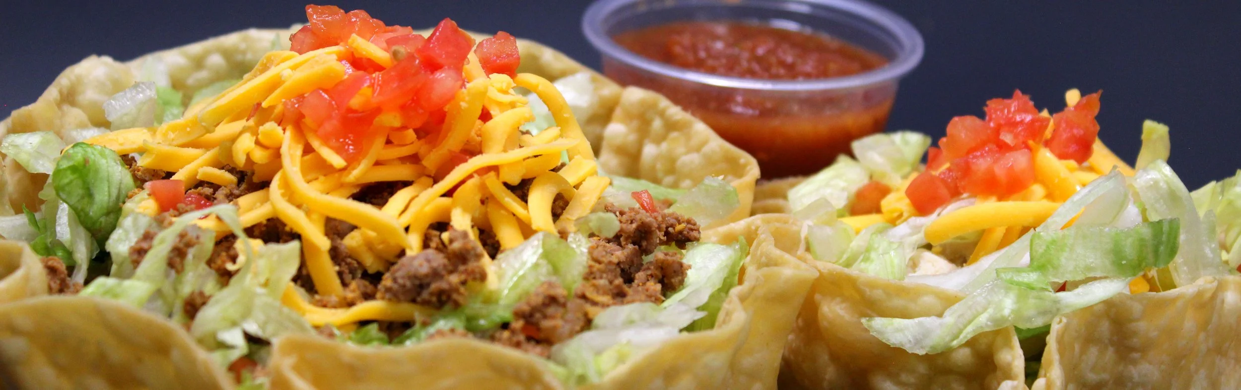 Close-up of seasoned taco salad with shredded cheese, lettuce, tomatoes, beef, in a fried tortilla bowl, with salsa in a small cup.