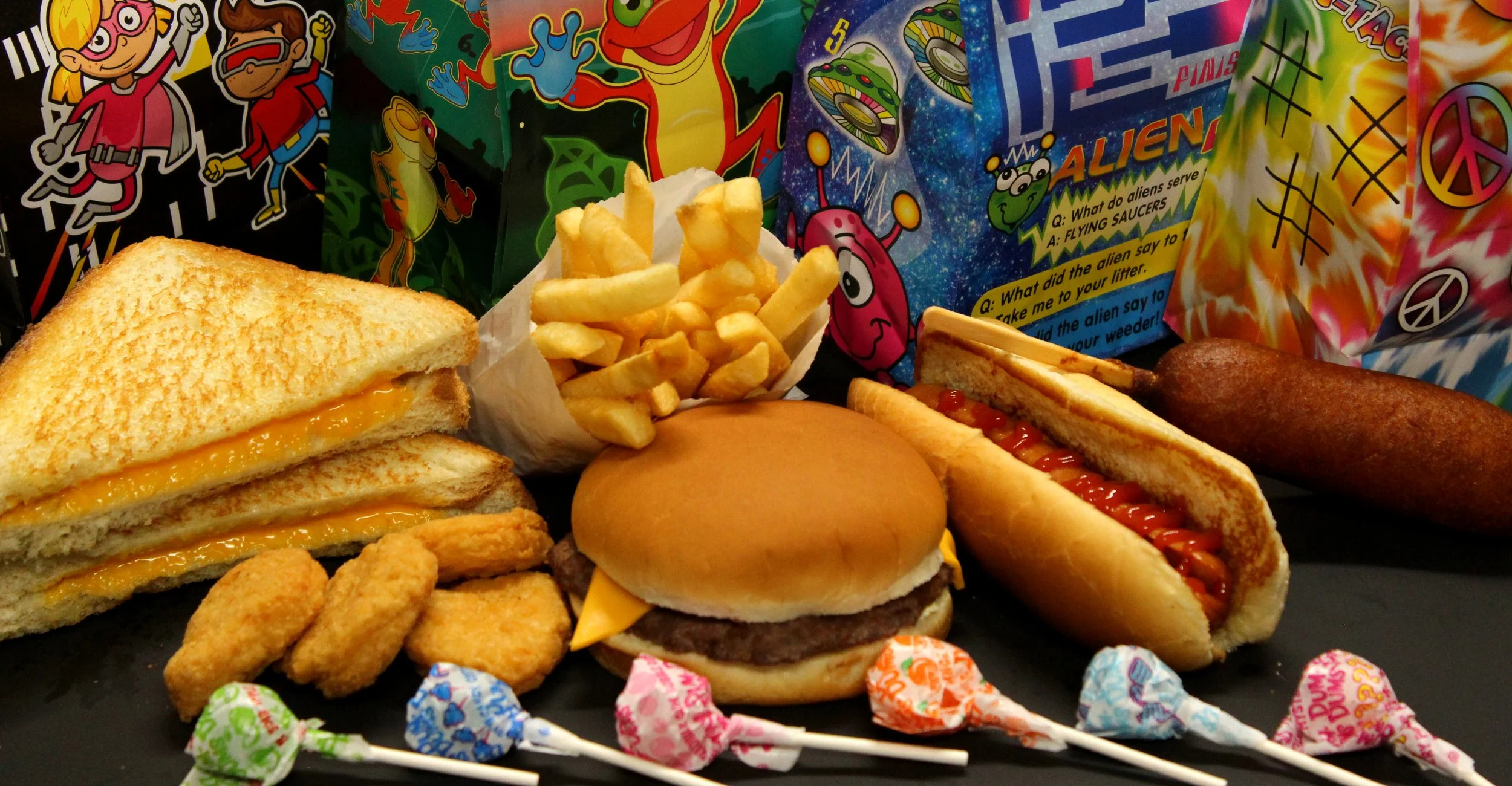 Fast food meal with grilled cheese sandwich, French fries, cheeseburger, hot dog, chicken nuggets, fried hot dog, and a corn dog, with colorful children's party favor bags in the background and small lollipops in the foreground.