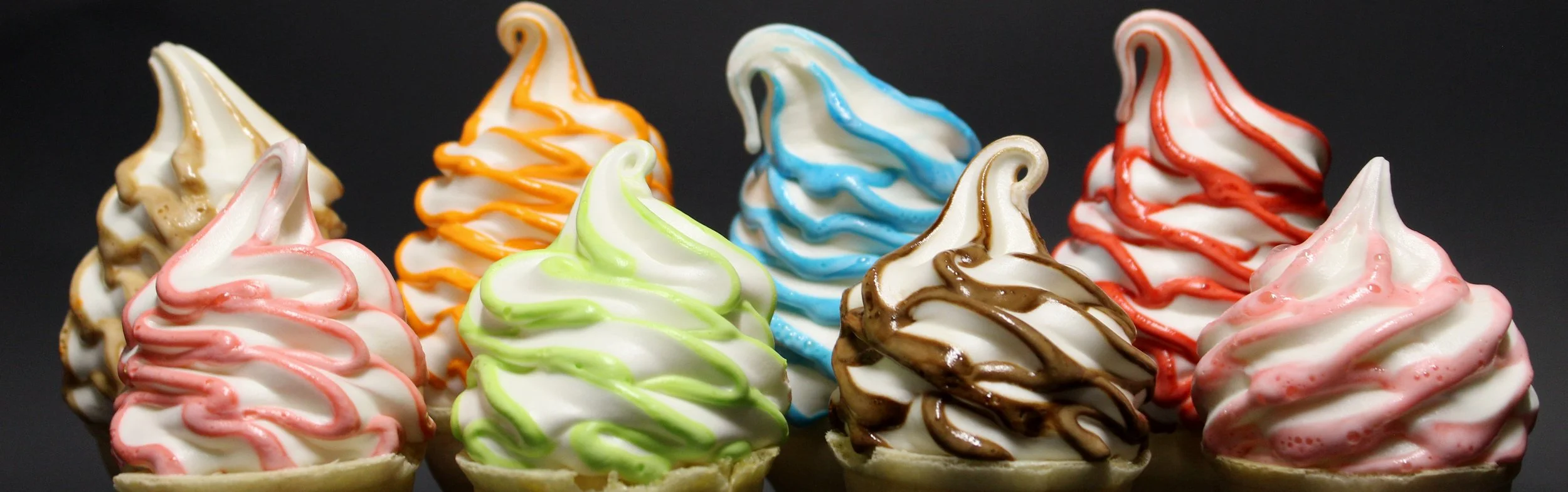 A row of colorful soft-serve ice cream cones topped with various brightly colored swirls of syrup, set against a black background.