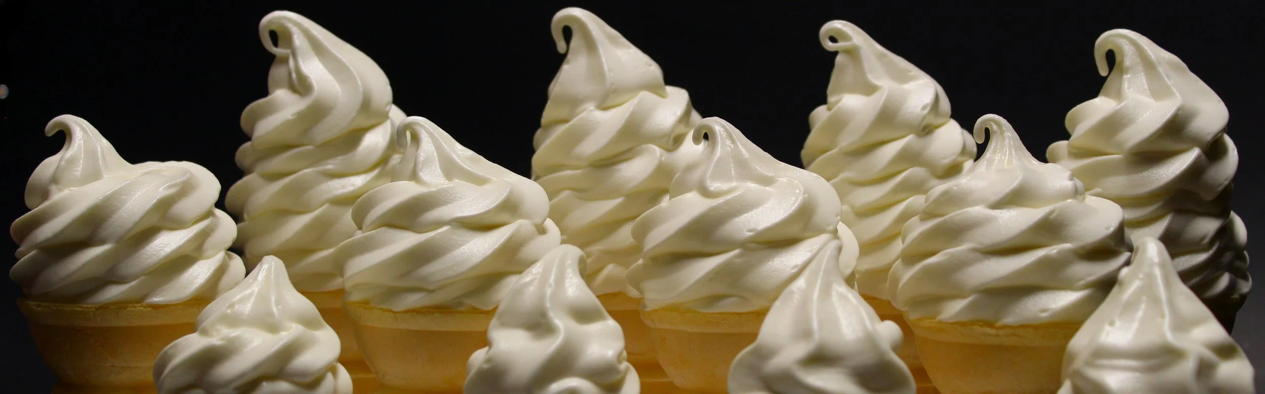 Multiple vanilla ice cream cones with swirled peaks in a row against a dark background.