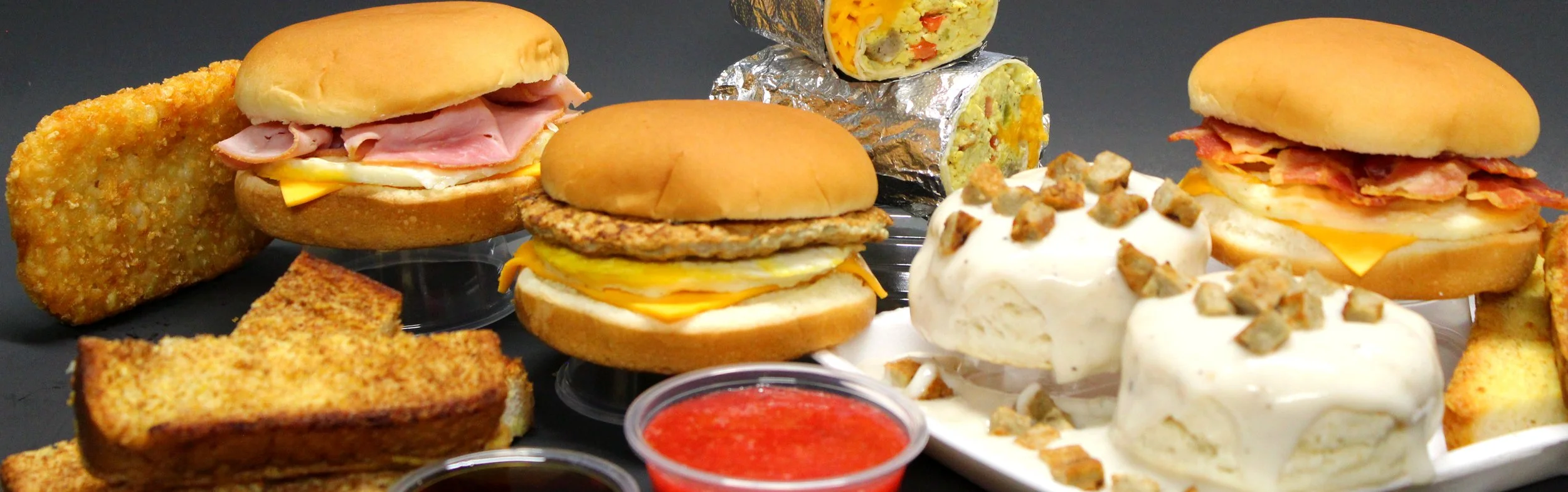 An assortment of breakfast sandwiches, biscuits and gravy, French toast slices, breakfast burrito wraps, hashbrowns, and small cups of dipping sauces on a black background.