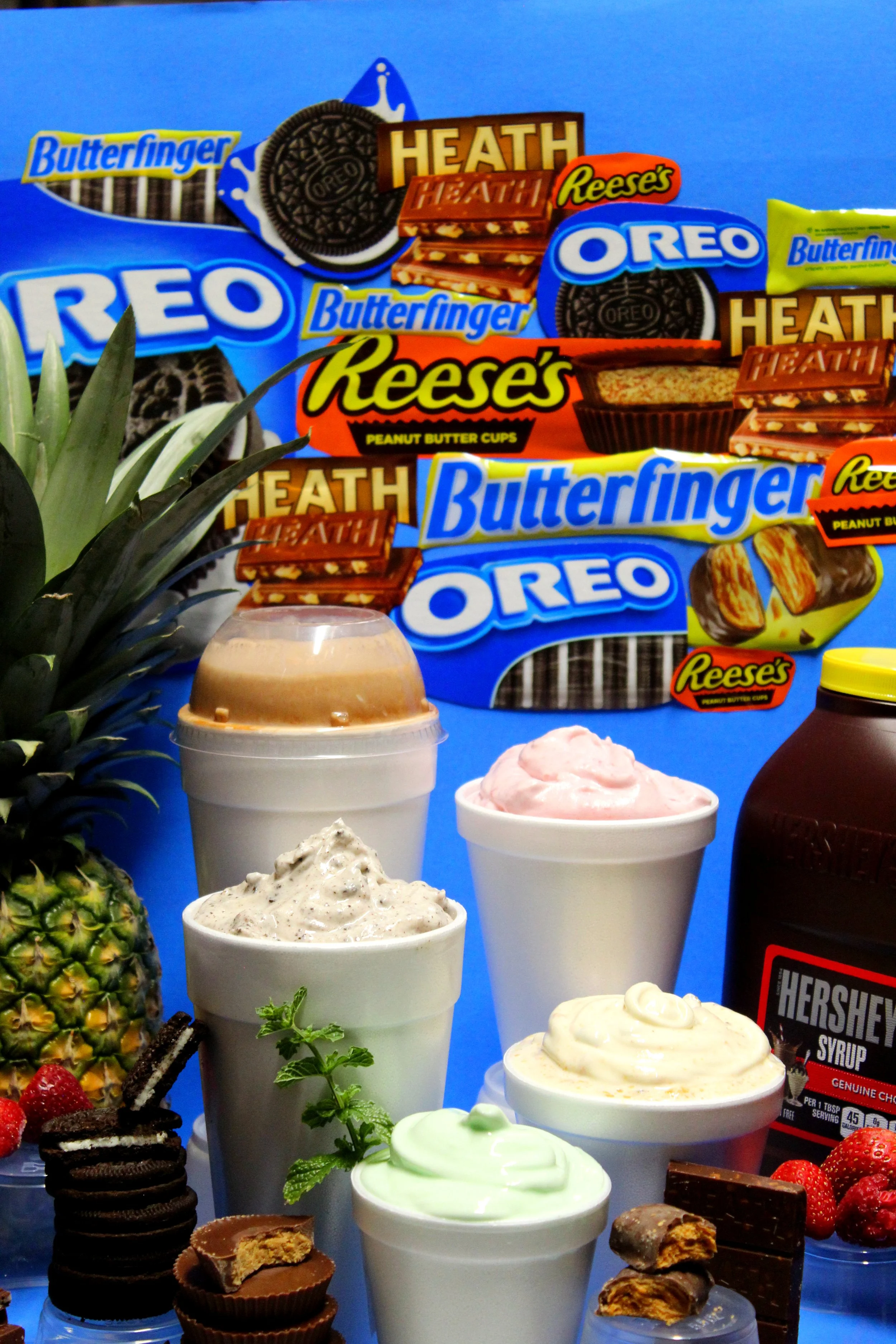 Assorted ice cream cups and desserts displayed with fruit, cookies, and a bottle of chocolate syrup in front of a colorful collage of various Oreo and Reese's snack packages.
