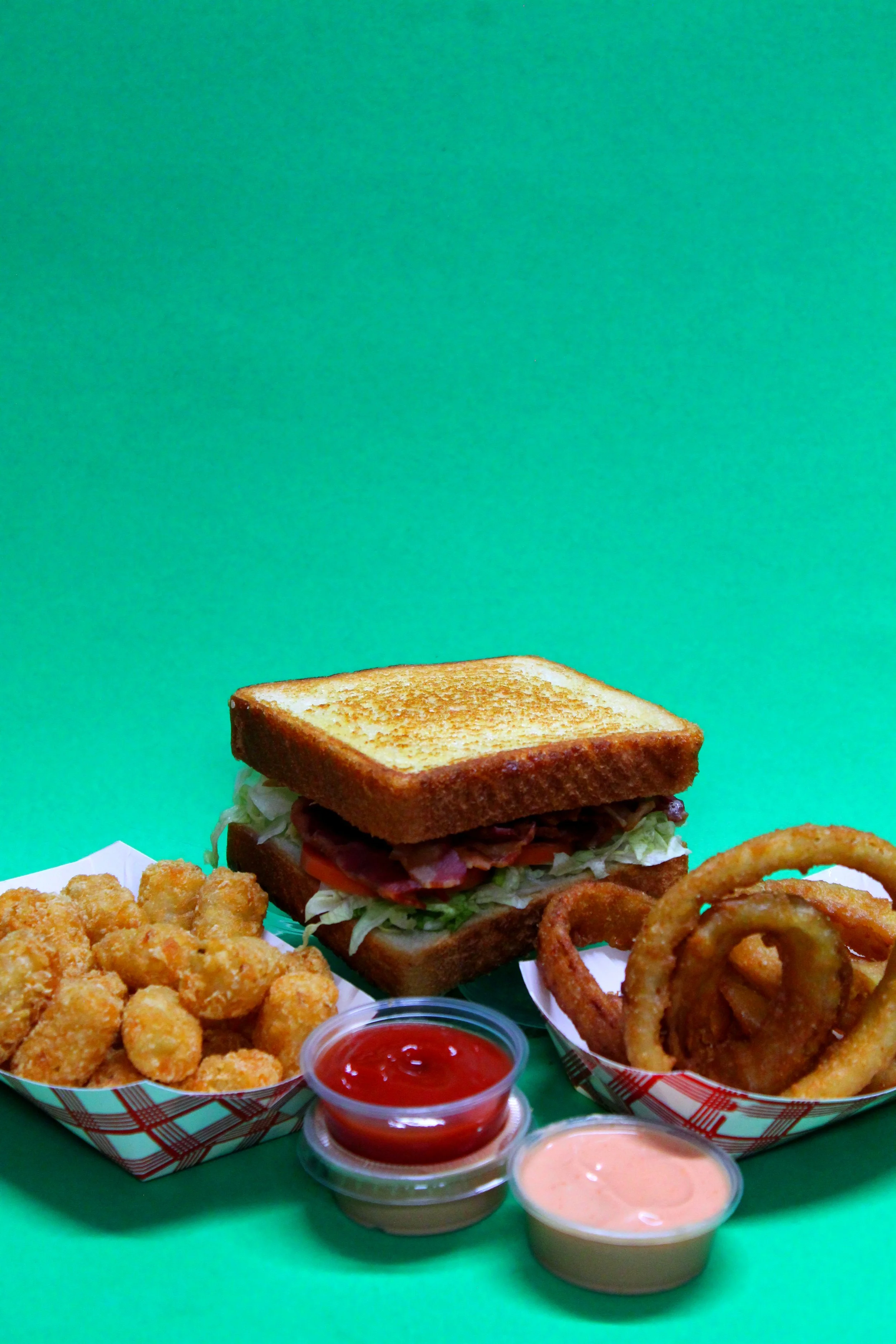 A plate of potato tots, a sandwich with toasted bread, bacon, lettuce, and tomato, onion rings, and two dipping sauces on a green background.