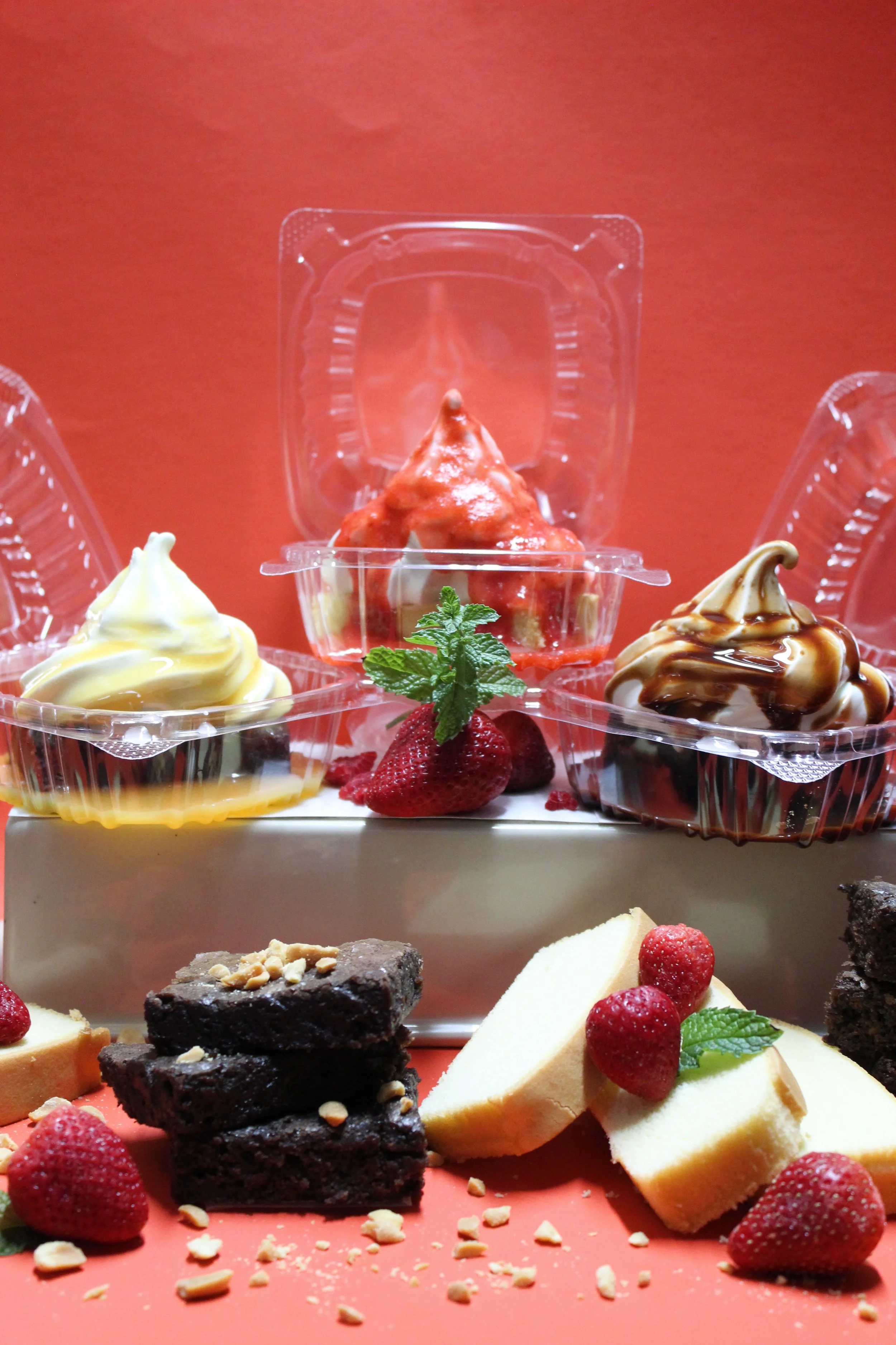 An assortment of chocolate brownies and slices of pound cake arranged with three servings of soft-serve ice cream topped with caramel, strawberry, and chocolate sauces on a white tray against a red background.