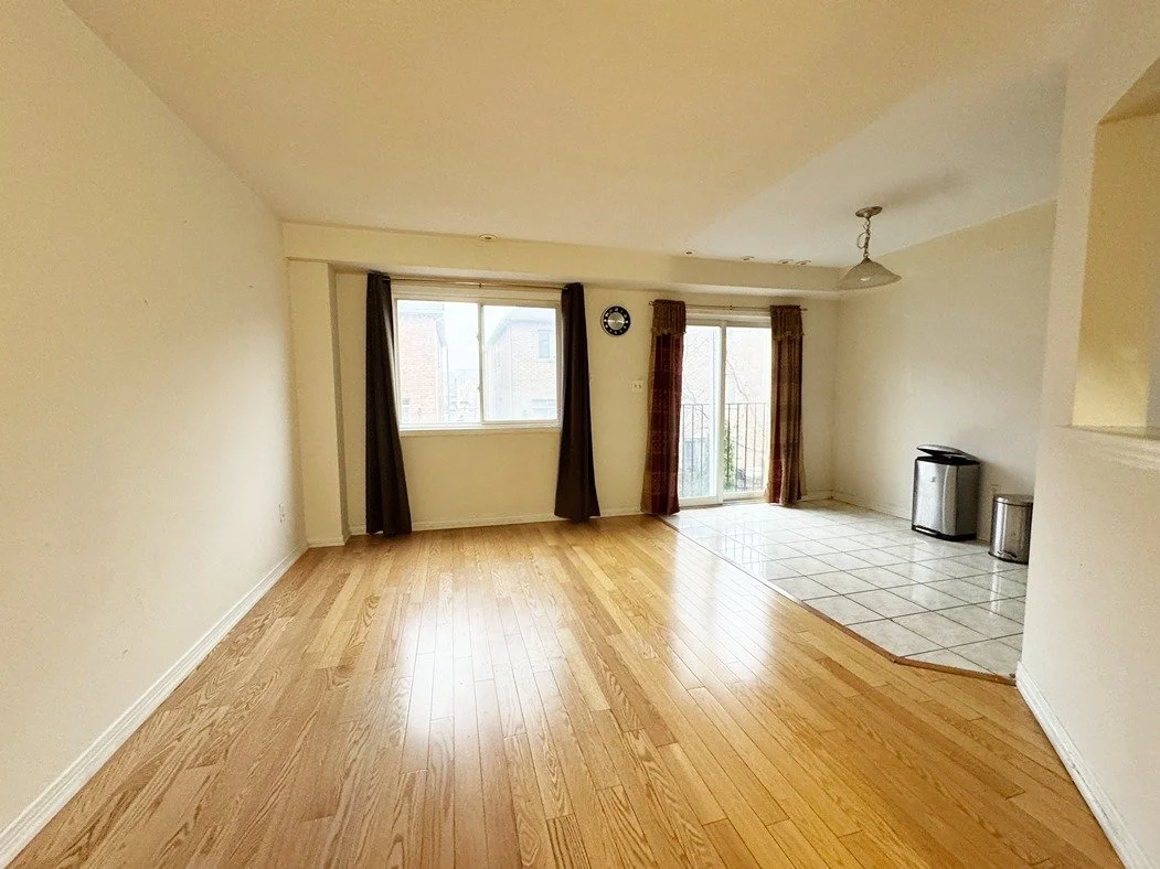 Empty living room with hardwood flooring, large window, sliding glass door with curtains, and a tiled area with a trash bin and mini fridge.