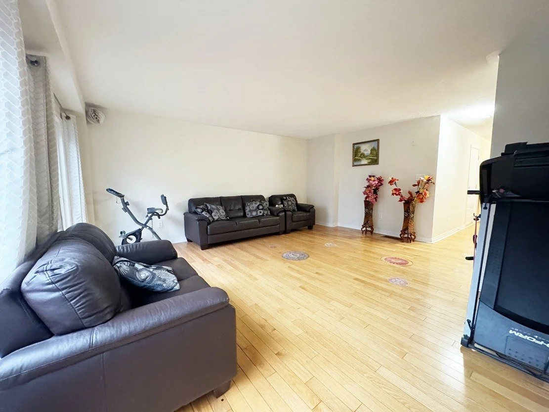 Living room with two black leather sofas, a stationary exercise bike, two tall decorative vases with pink and red flowers, and a framed landscape painting on the wall.