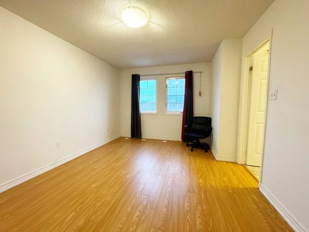 Empty living room with white walls, wooden flooring, two windows with dark curtains, a black chair, and an open door leading to another room.