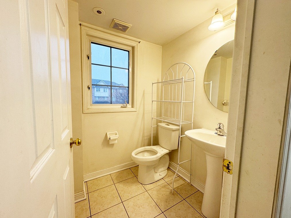 Small bathroom with a window, toilet, pedestal sink, round mirror, wall-mounted light fixtures, and a white metal shelving unit.