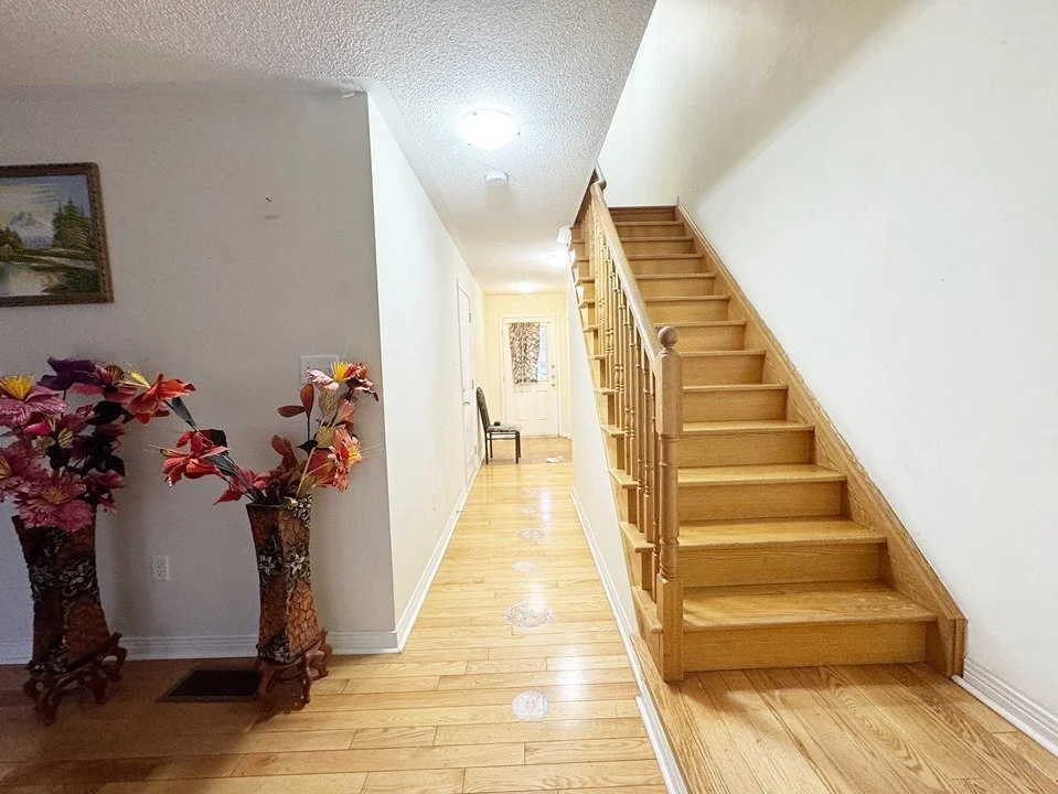A hallway with wooden flooring, a staircase with wooden steps and railing, two decorative vases with artificial flowers, a framed picture on the wall, a chair at the end of the hall, and a closed door with a curtain covering the window.