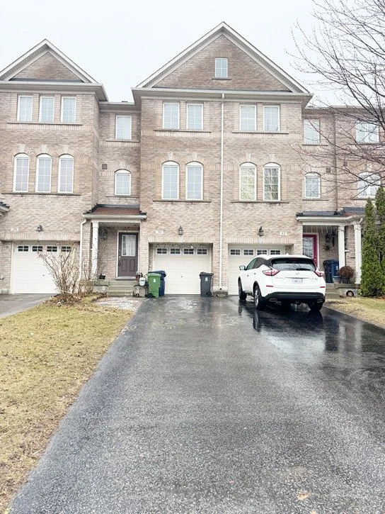 Row of three attached townhomes with brick facades, each with a garage and driveway. The middle townhome has a white SUV parked on its driveway. Overcast sky, wet pavement, and leafless trees.