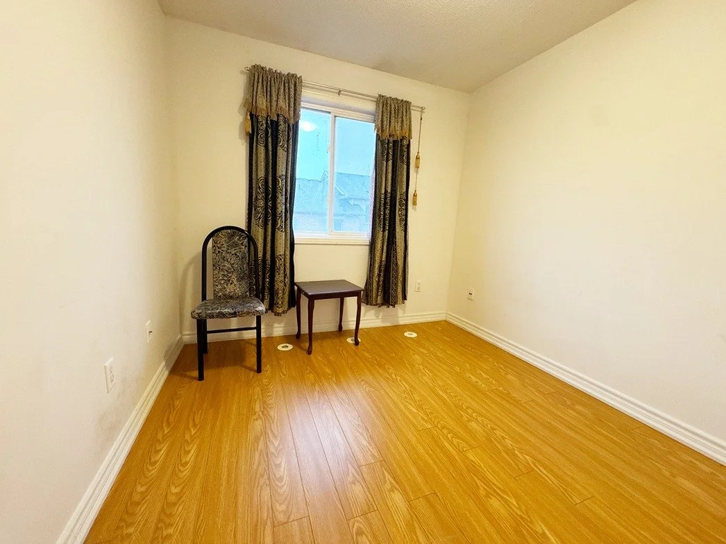 Empty room with hardwood floors, a window with patterned curtains, a single chair, and a small table.