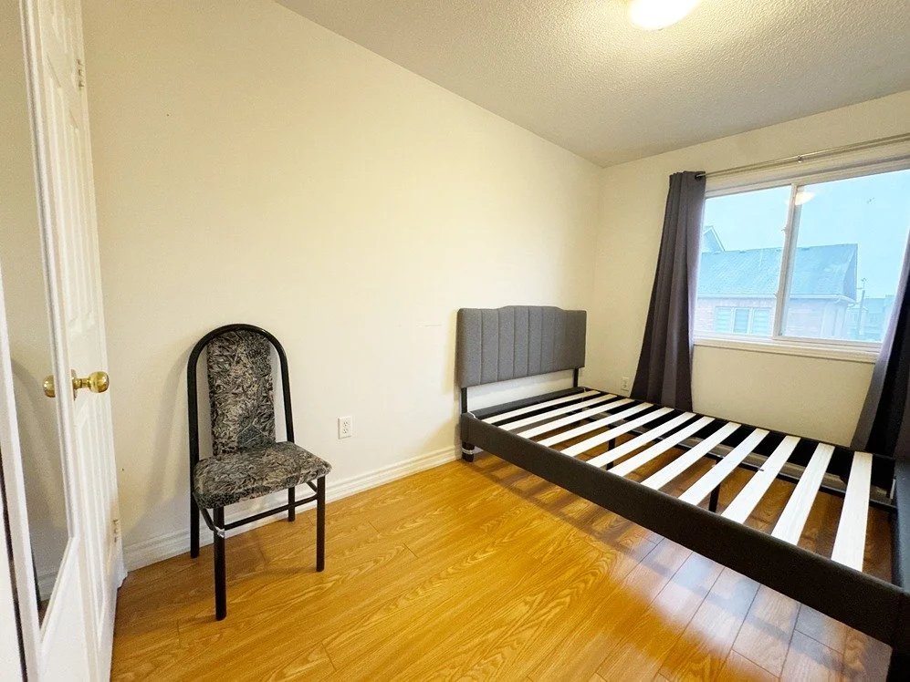 Empty bedroom with a bed frame, a patterned chair, a window with curtains, and wood flooring.