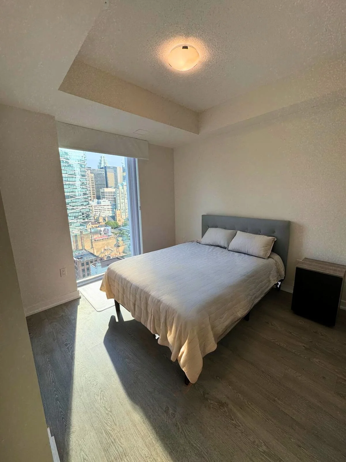 A sunlit bedroom with a large bed, light-colored bedding, and two pillows. There is a window showing a cityscape of high-rise buildings. The ceiling has a light fixture, and there's a small nightstand beside the bed.