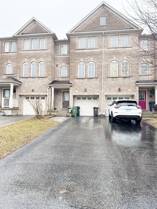 A row of three-story townhouses with brick facades, parked cars, and trash bins in front. The driveways are wet, suggesting recent rain, and there are small patches of grass beside the driveways.