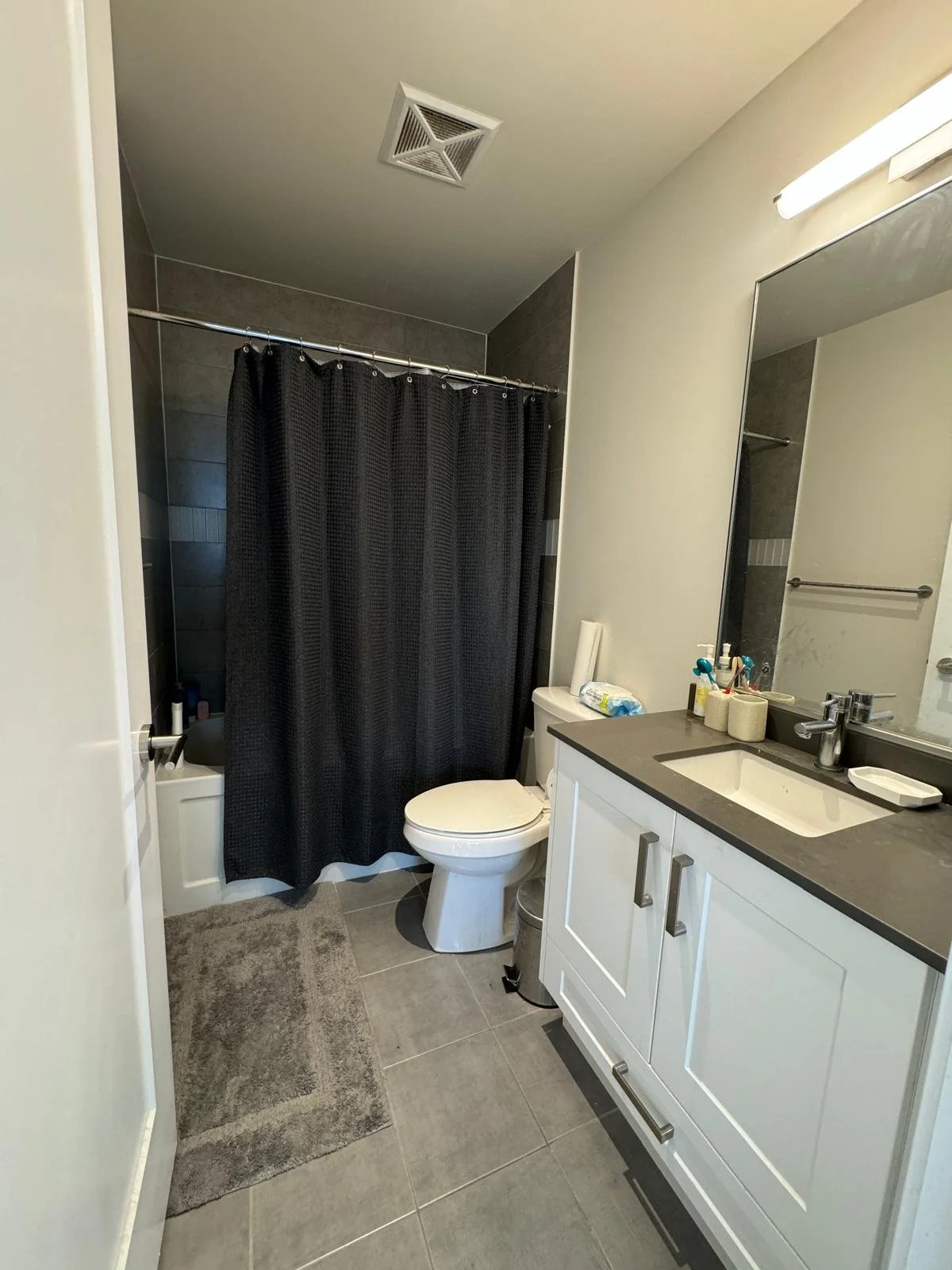 Small bathroom with a toilet, vanity with a sink, mirror, and a shower with a black curtain. Gray tiled floor and dark wall tiles in the shower area. Bathroom items on the vanity and a trash can.