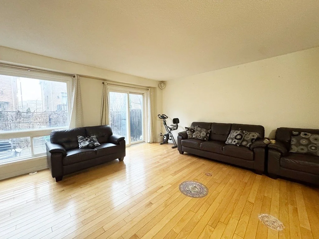 Living room with hardwood floors, three black leather sofas with patterned pillows, large windows with curtains, a stationary bike, and a sliding glass door leading outside.