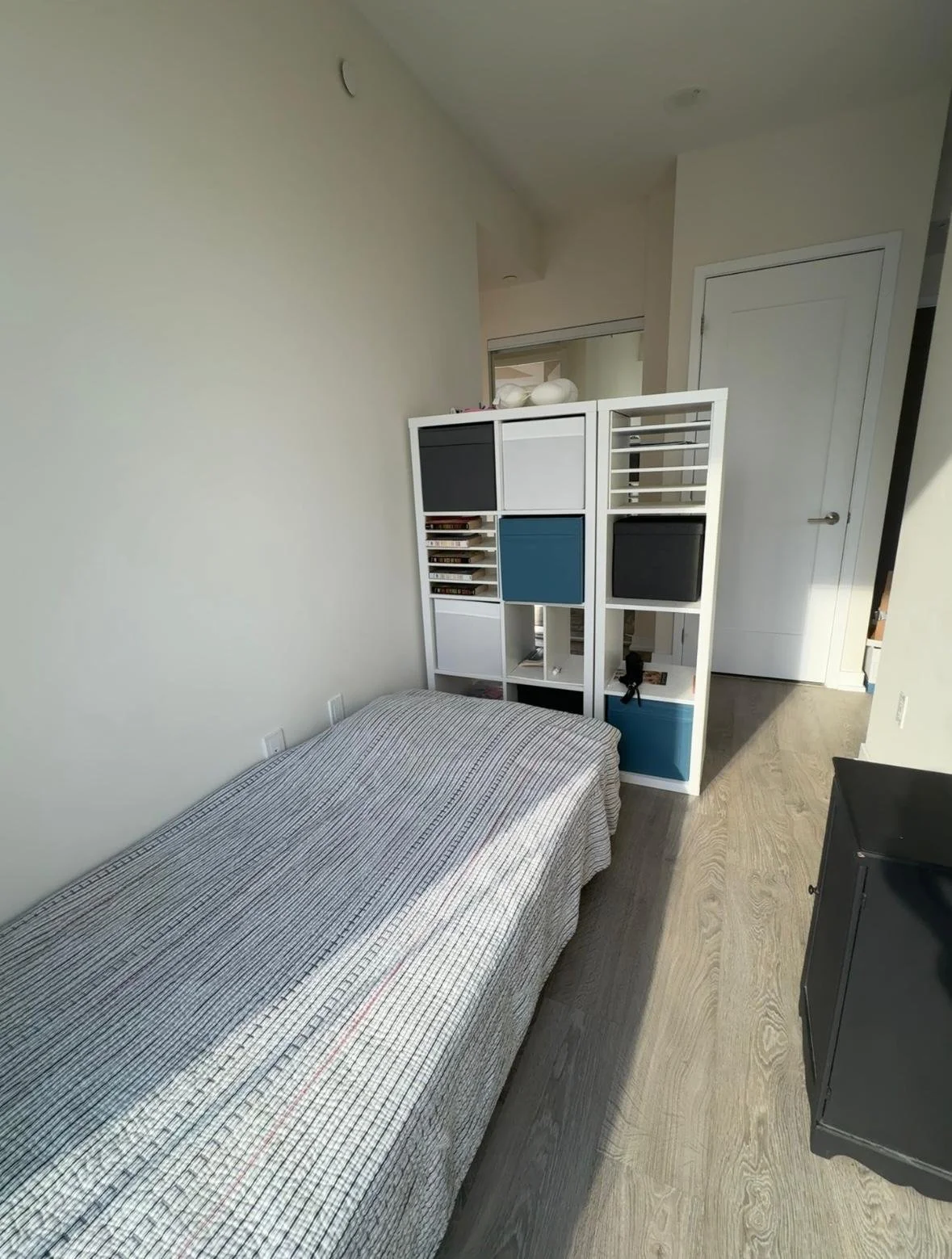 A small bedroom with a single bed covered in a striped blanket. There is a white shelving unit behind the bed with various compartments, some containing DVDs and others with decorative items. The room has a light-colored hardwood floor, a dark small