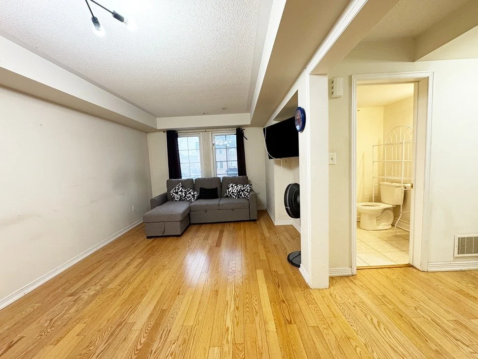 Living room with hardwood floors, gray sectional sofa with patterned pillows, a wall-mounted TV, a window with black curtains, and a visible bathroom with a toilet and a metal rack.