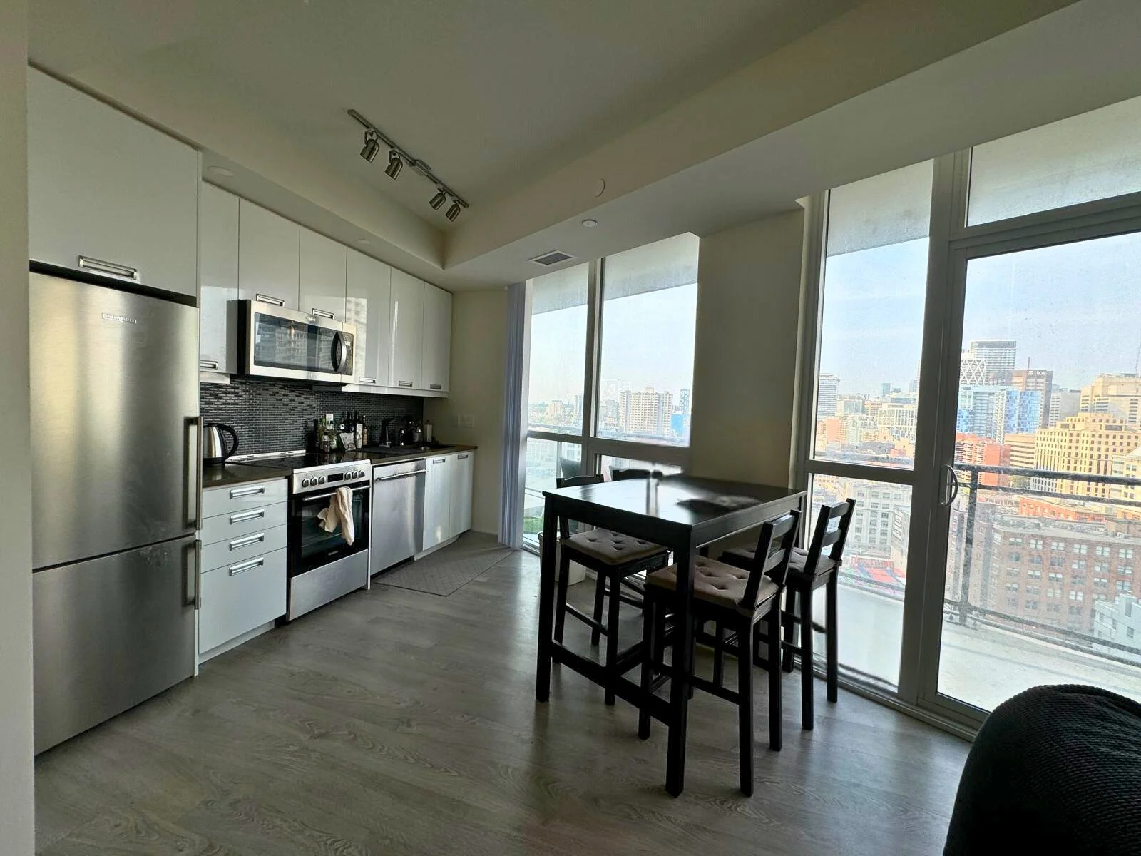 Modern kitchen and dining area with large windows showing a city view, white cabinets, stainless steel appliances, black backsplash, a small dining table with four chairs.