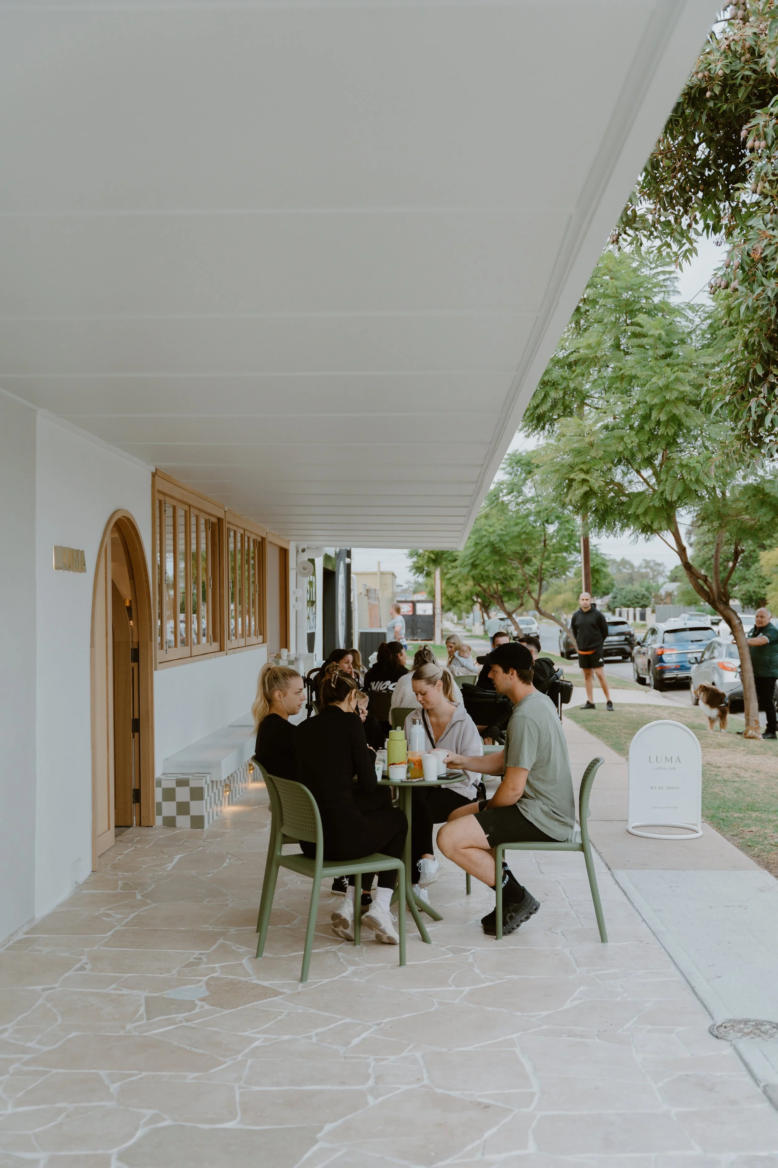 People sitting at tables outside a cafe on a sidewalk, with trees and parked cars in the background.