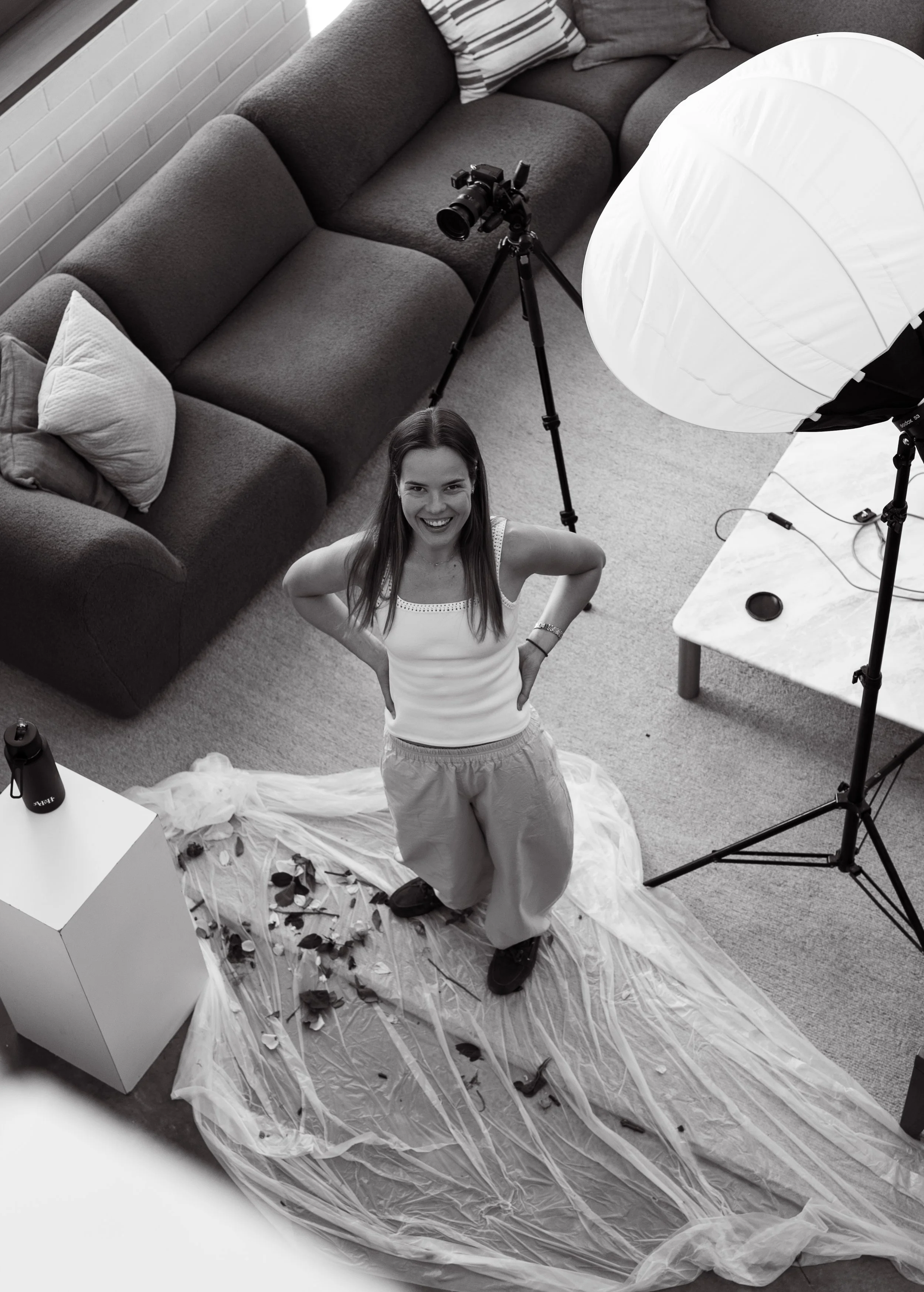 A woman smiling and standing with her hands on her hips in a room, with studio lighting equipment, a camera on a tripod, and a sofa in the background, as part of a photo shoot setup.