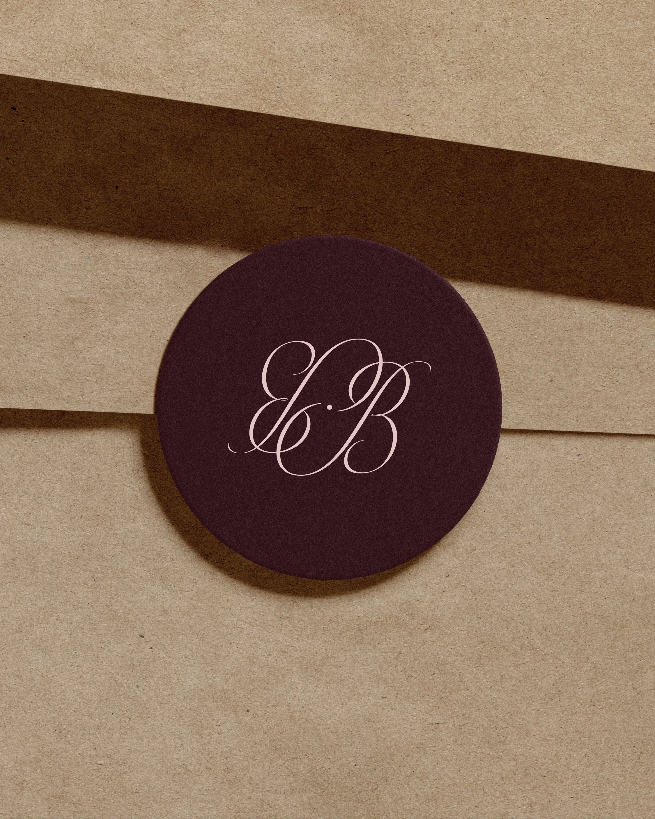 Close-up of a round, dark purple object with white cursive initials 'B B' on it, placed on a beige textured surface with shadows.