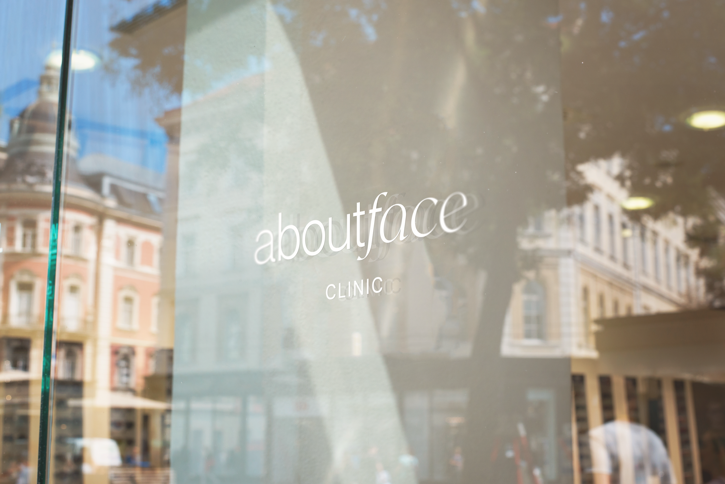 A glass window with the words 'about face CLINIC' printed on it, reflecting buildings and trees from outside.