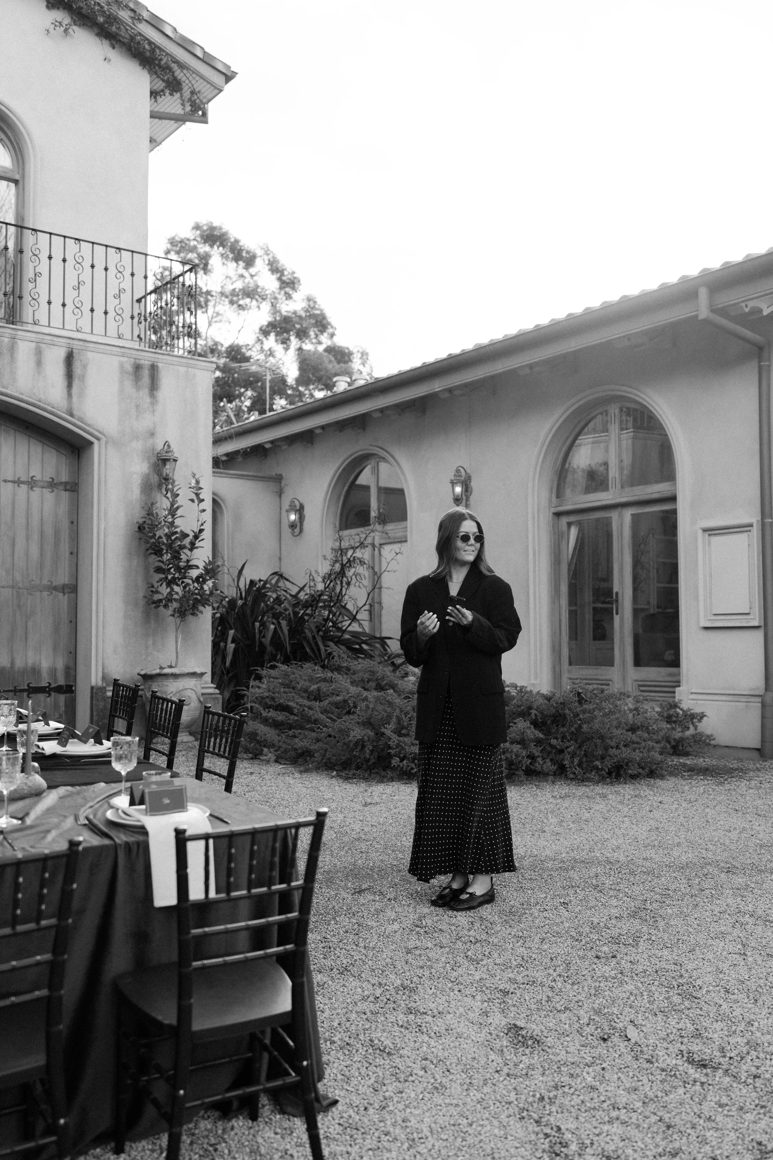 A woman with long hair wearing sunglasses, a dark blazer, and polka dot maxi skirt, standing outdoors in a courtyard with a table set for a meal, in front of a light-colored house with arched windows, bushes, and trees.