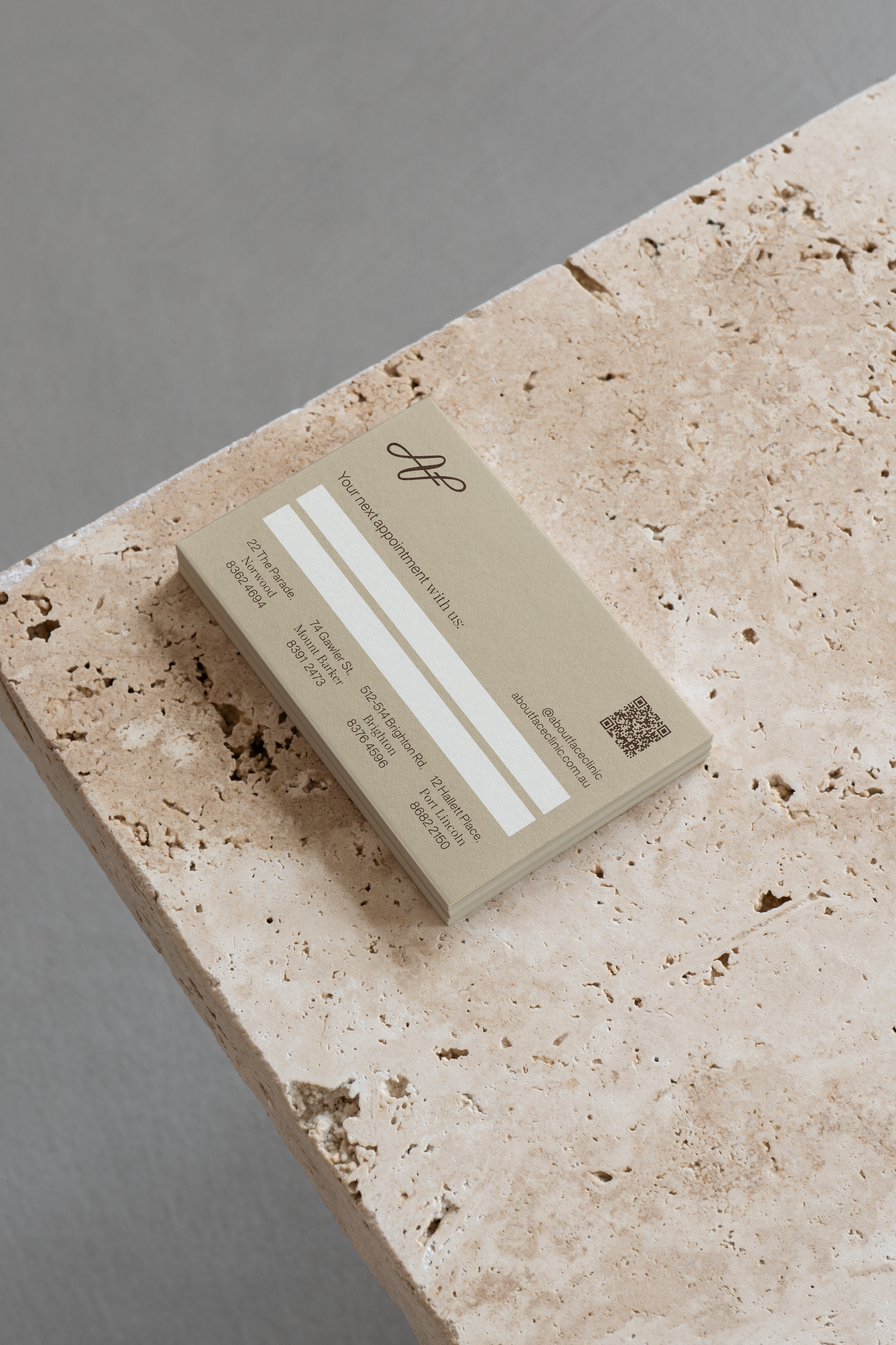 Business card placed on a beige, textured stone surface. The card has black text and a QR code.