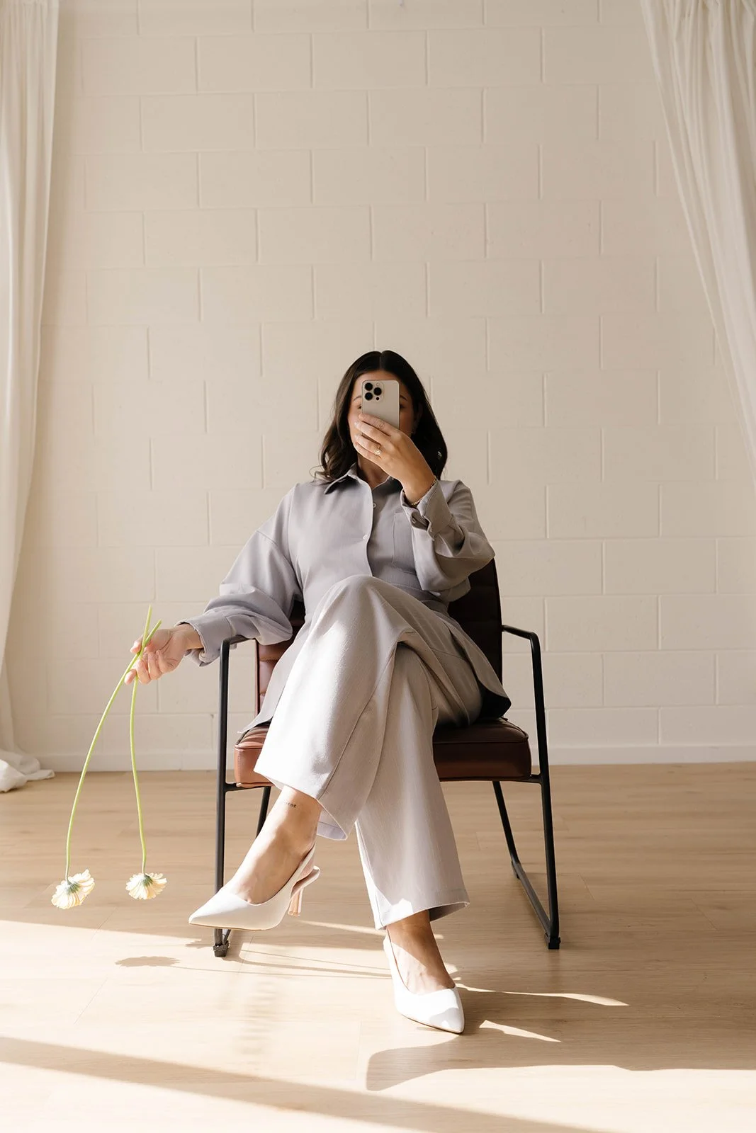 A woman sitting on a chair in a room with a white brick wall, wearing a light gray oversized shirt and wide-leg trousers, holding flowers, and taking a mirror selfie with her phone.